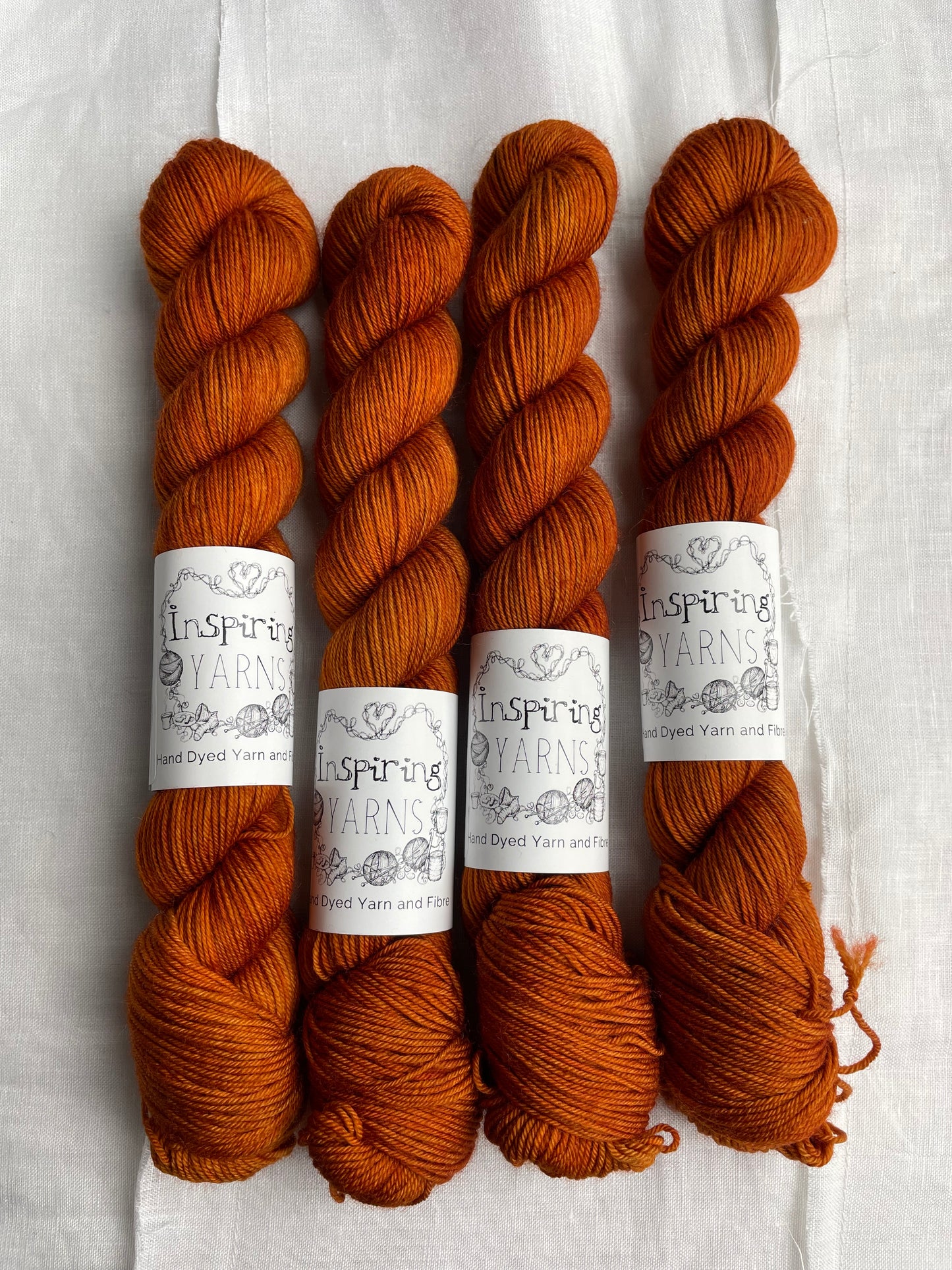 Four tonal burnt orange coloured skeins of hand dyed Merino Smooth Sock yarn branded Inspiring Yarns on white linen fabric.
