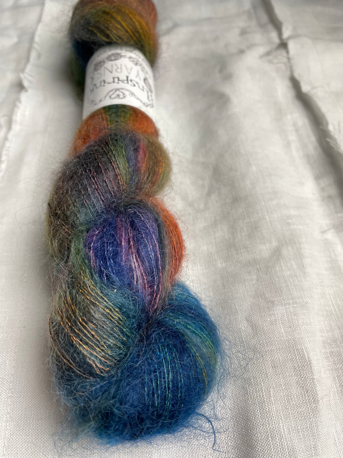 A skein of variegated blue and multi coloured kid mohair silk lace yarn labelled Inspiring Yarns on a white linen background.