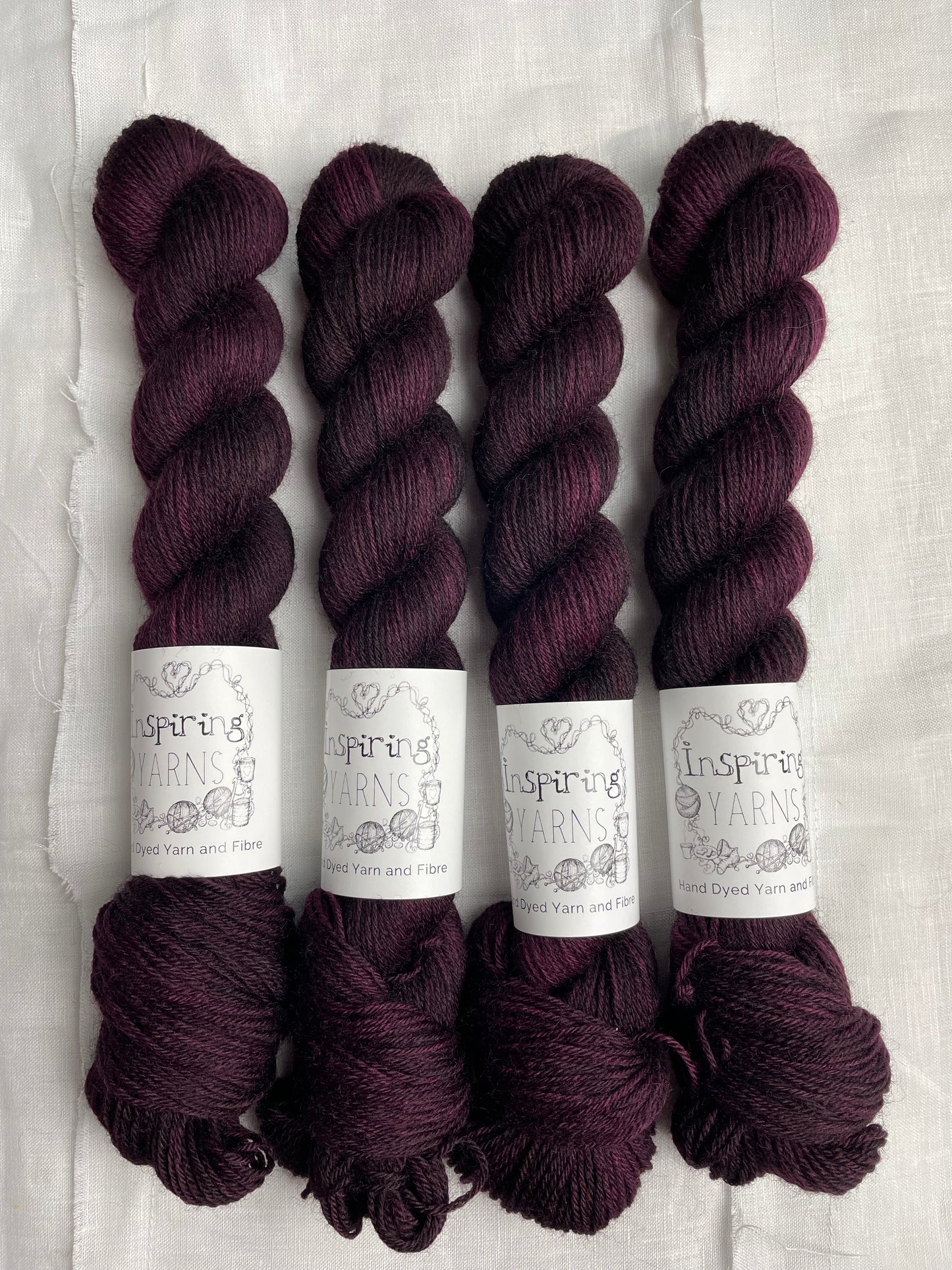 Four skeins of 50 gram dark burgundy coloured Highland 4Ply labelled Inspiring Yarns on a white linen background.