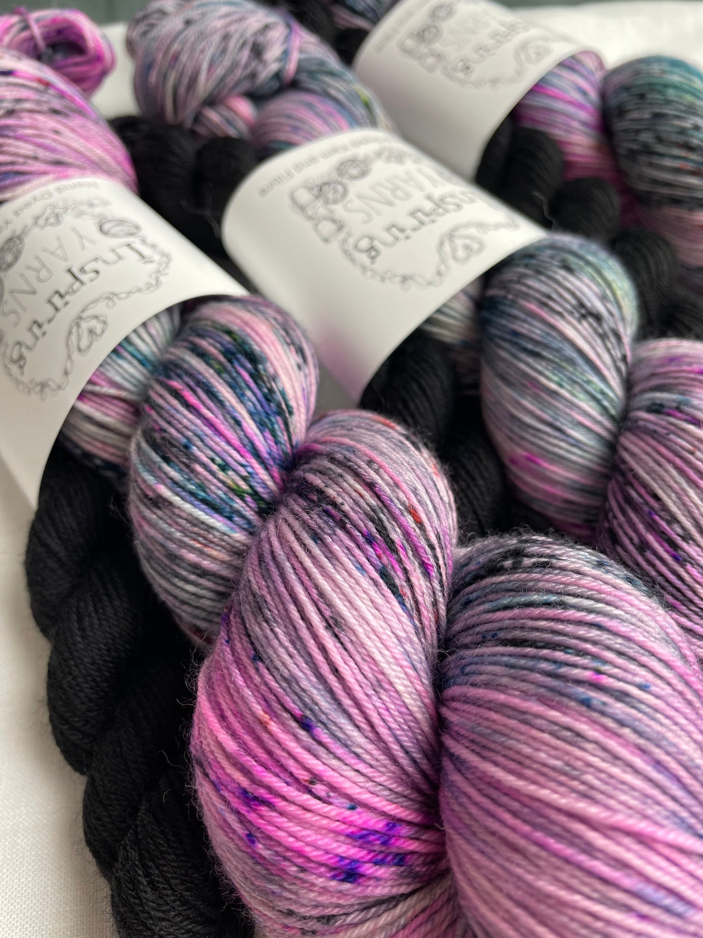 Close up of three sock sets made of a skein of pink black and teal speckled hand dyed yarn with a contrasting black mini skein, labelled Inspiring Yarns and laid on a white linen cloth.