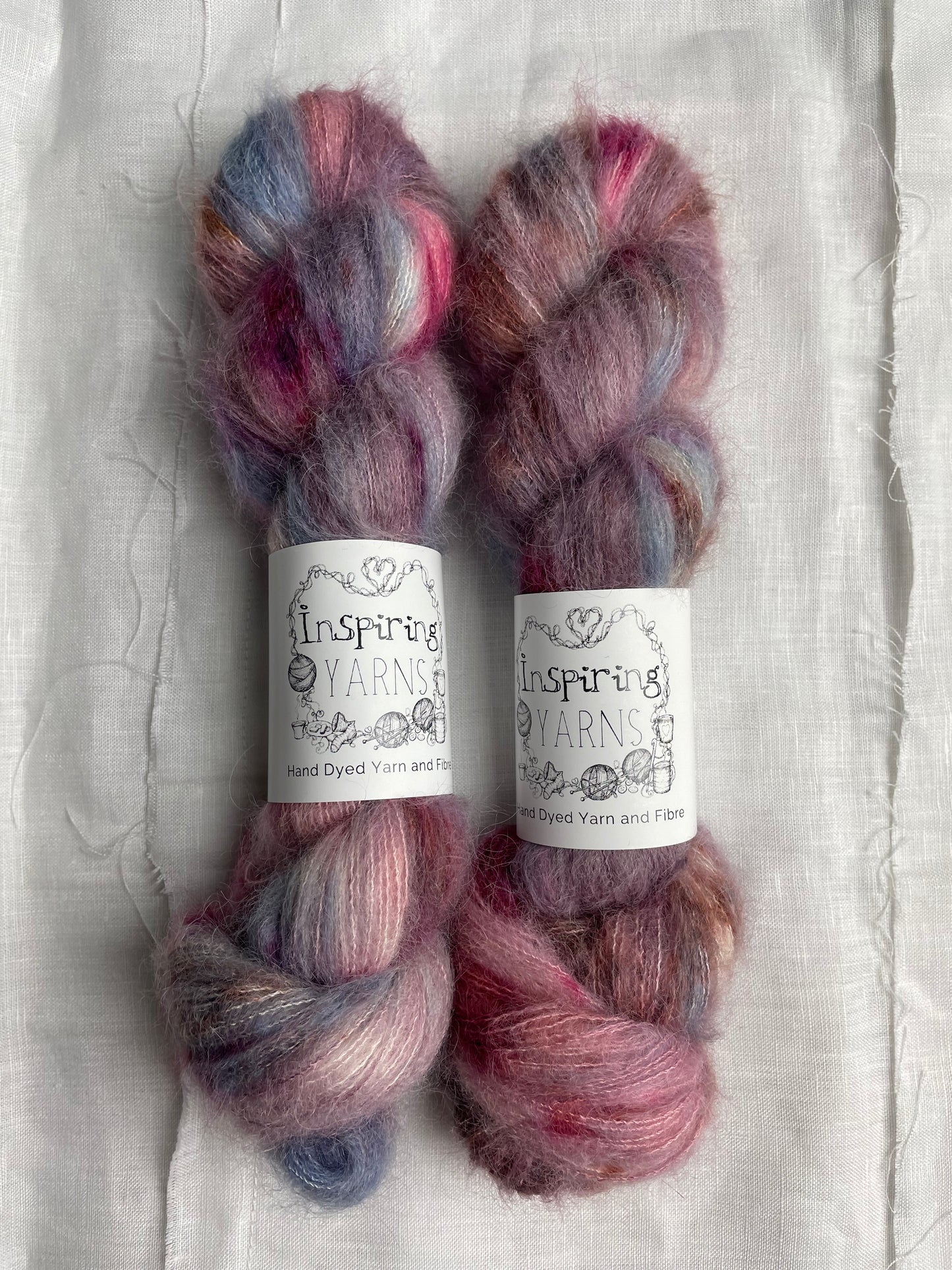 Two skeins of brushed Suri alpaca in shades of pink and blue labelled Inspiring Yarns on a white linen background.