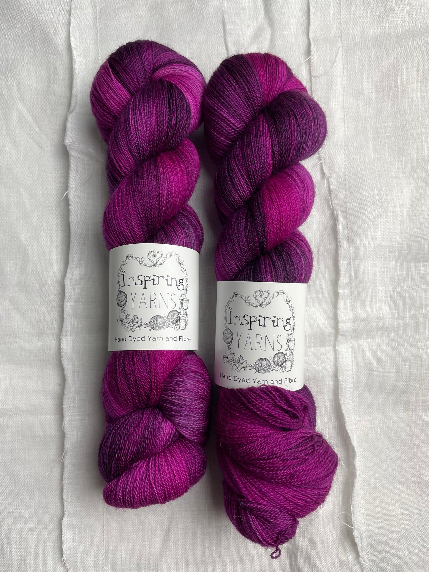 Two skeins of variegated pink plum coloured hand dyed Merino Lace yarn branded Inspiring Yarns on a white linen cloth.