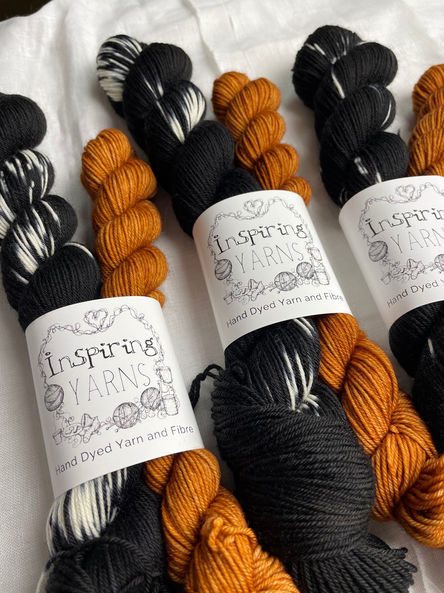 Close up of three sock sets made of a skein of black and white hand dyed yarn with a contrasting pumpkin orange mini skein, labelled Inspiring Yarns and laid on a white linen cloth.