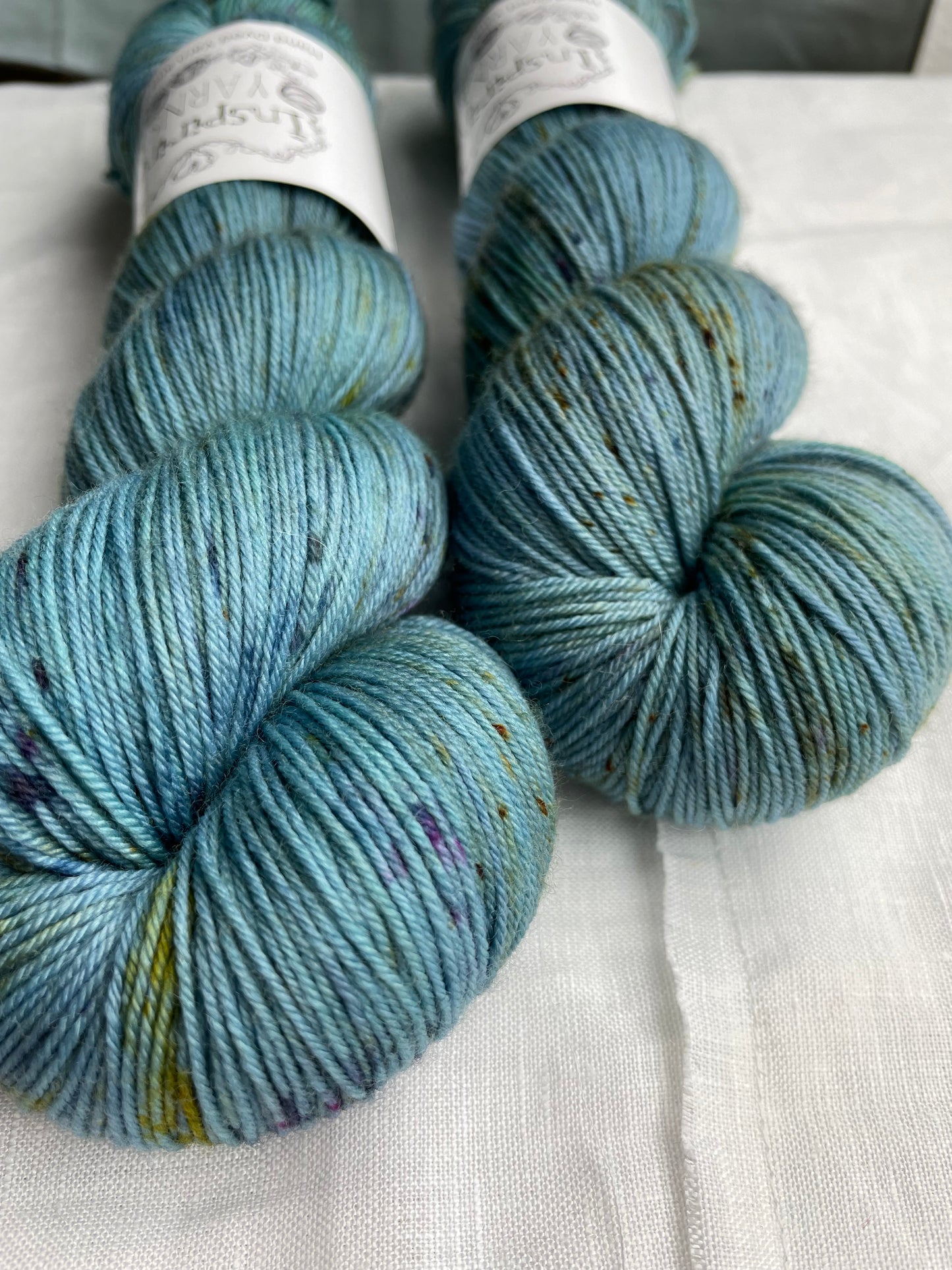 Close up of two speckled soft teal coloured skeins of Merino Smooth Sock yarn branded Inspiring Yarns on a white linen fabric background.