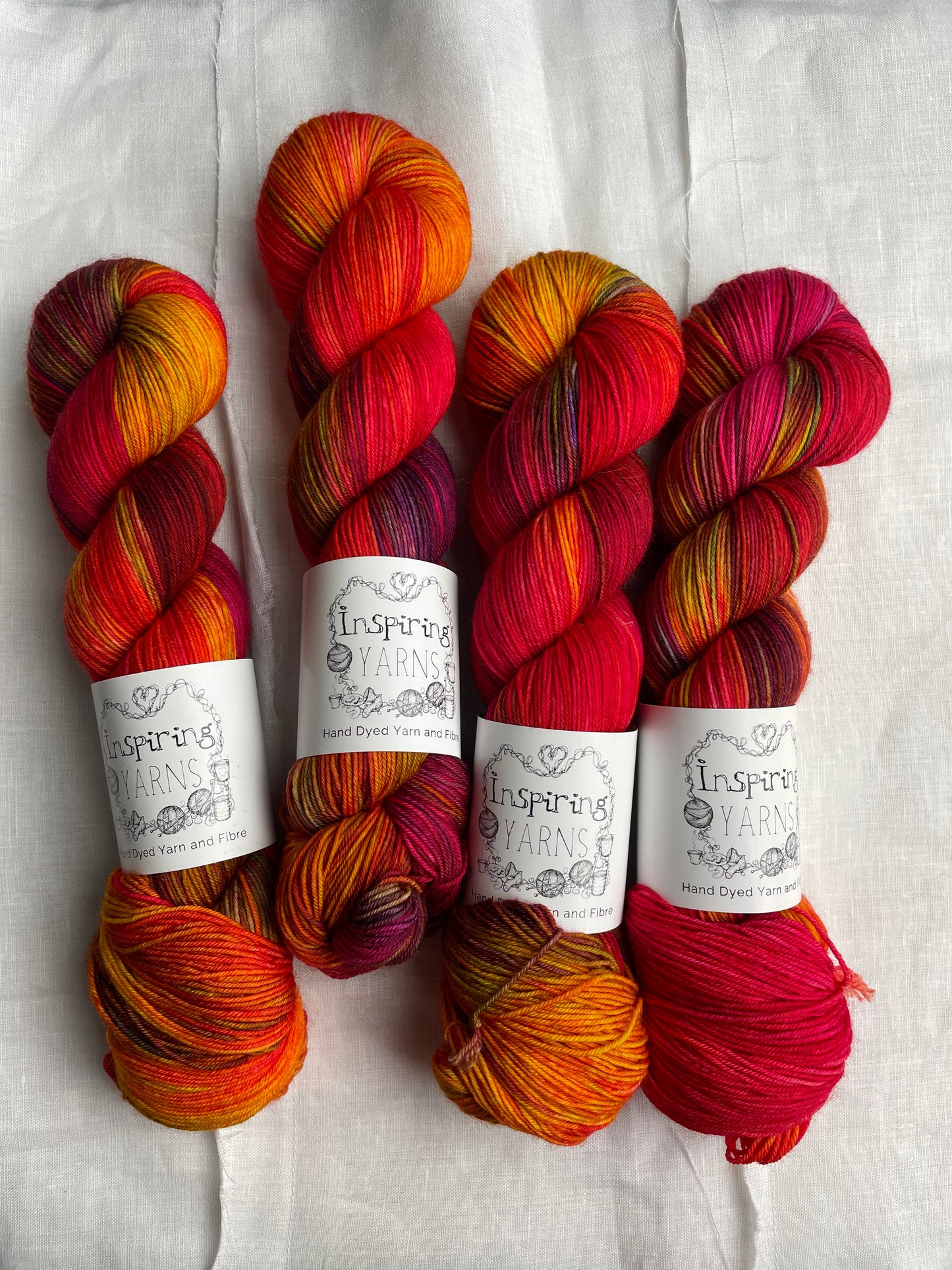 Four variegated red and multi coloured skeins of Merino Smooth Sock yarn branded Inspiring Yarns on a white linen fabric background.
