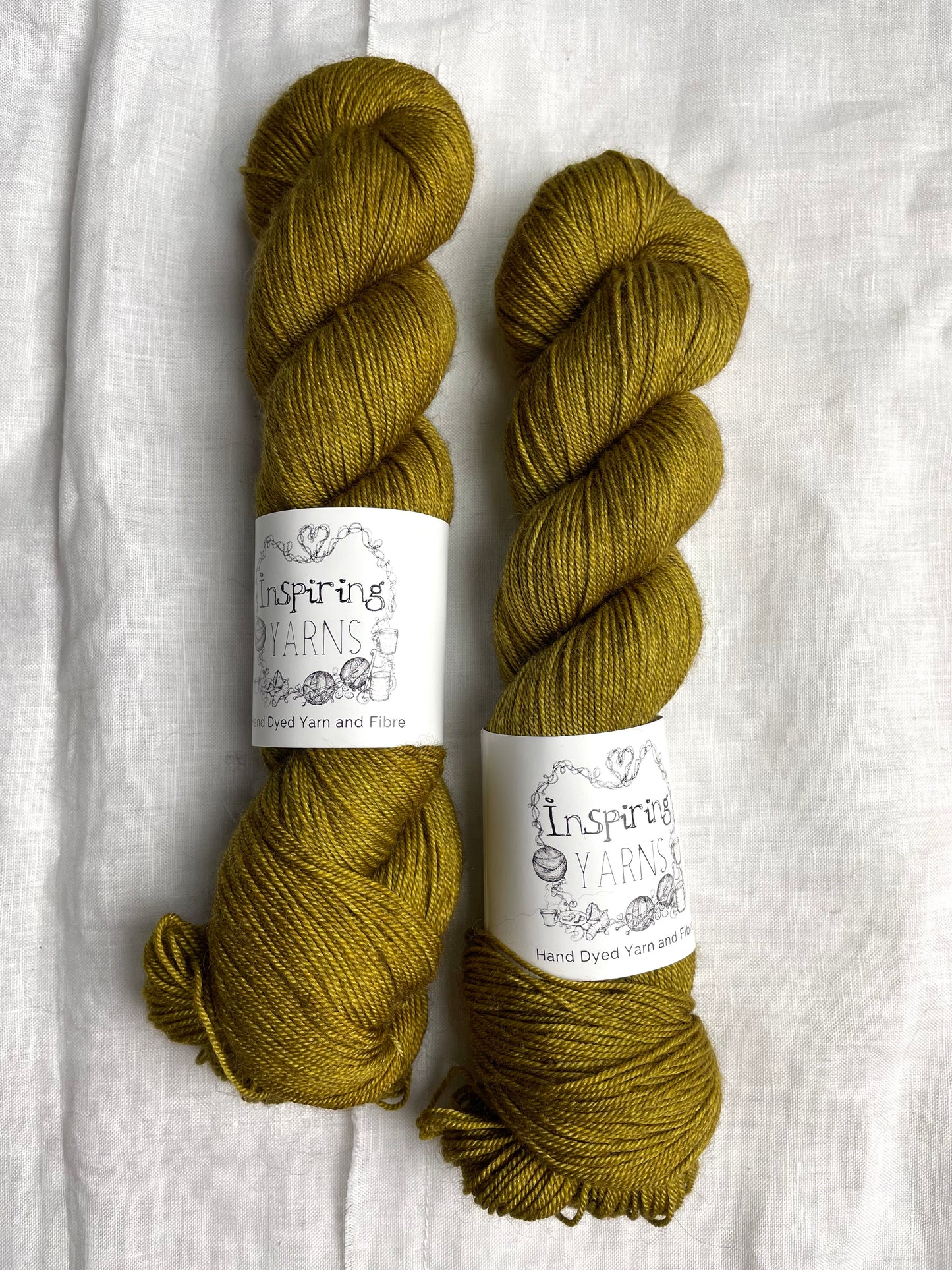 Two skeins of ochre yellow Yak Silk 4ply yarn labelled Inspiring Yarns on a white linen background.