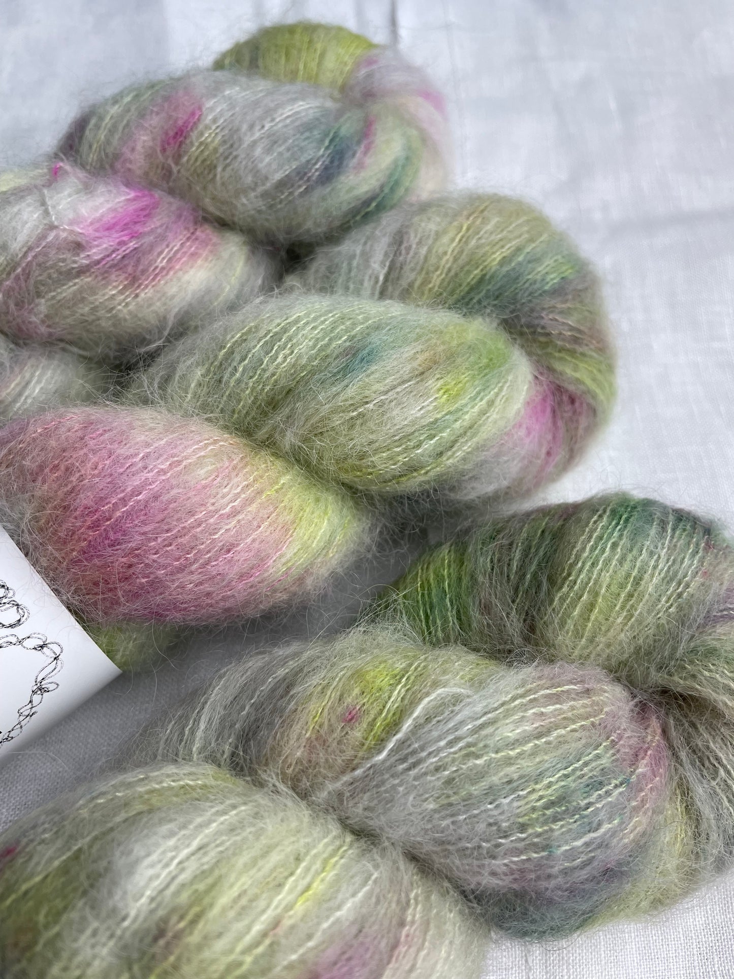 Close up of three skeins of green and pink speckled silver grey coloured hand dyed fluffy Alpaca Silk Lace yarn labelled Inspiring Yarns on a white linen background.