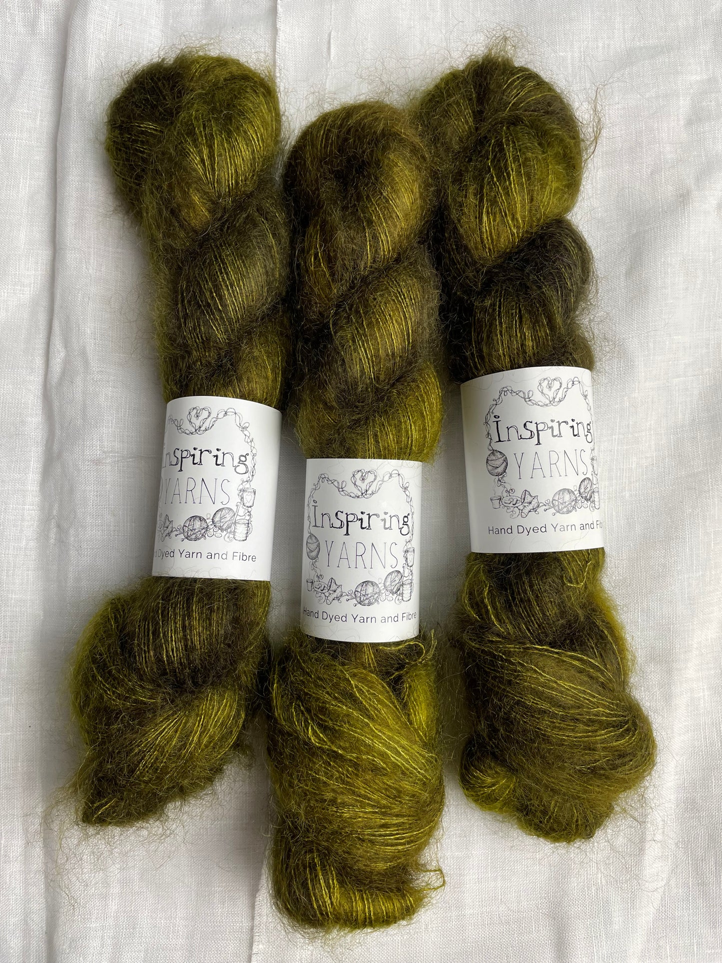 Three skeins of dark olive green mohair silk lace yarn labelled Inspiring Yarns on a white linen background.