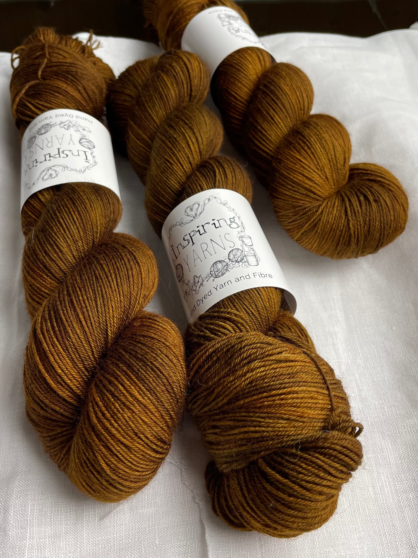 Three skeins of Kelp, a bronze tonal coloured Merino Smooth Sock, labelled Inspiring Yarns on a white linen background.