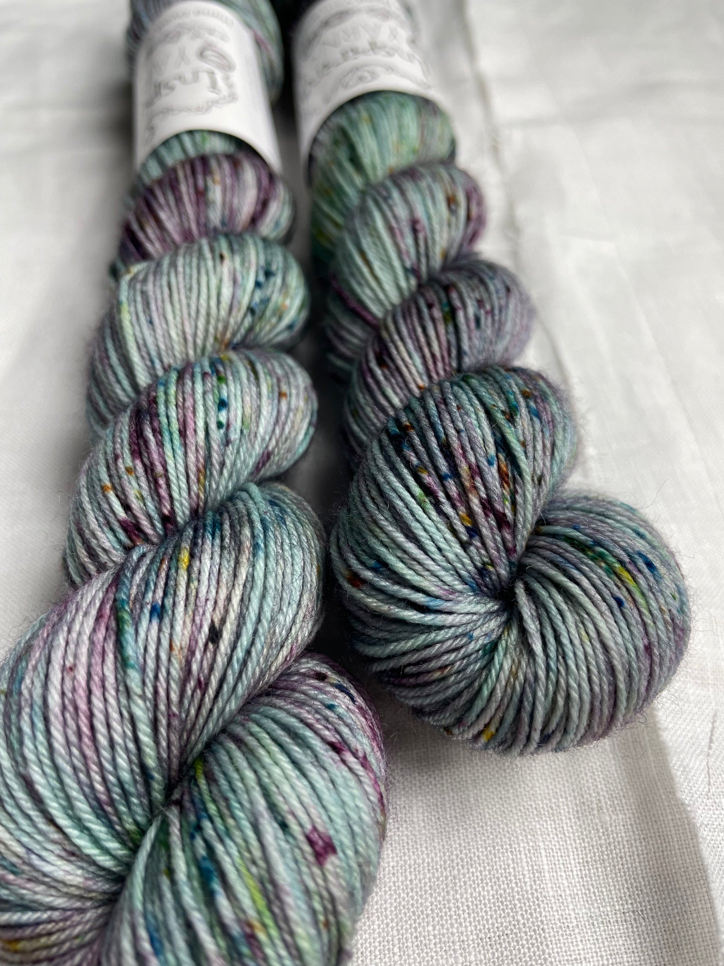 Two skeins of merino smooth sock yarn in a light teal colour with coloured speckles labelled Inspiring Yarns on a white linen background.