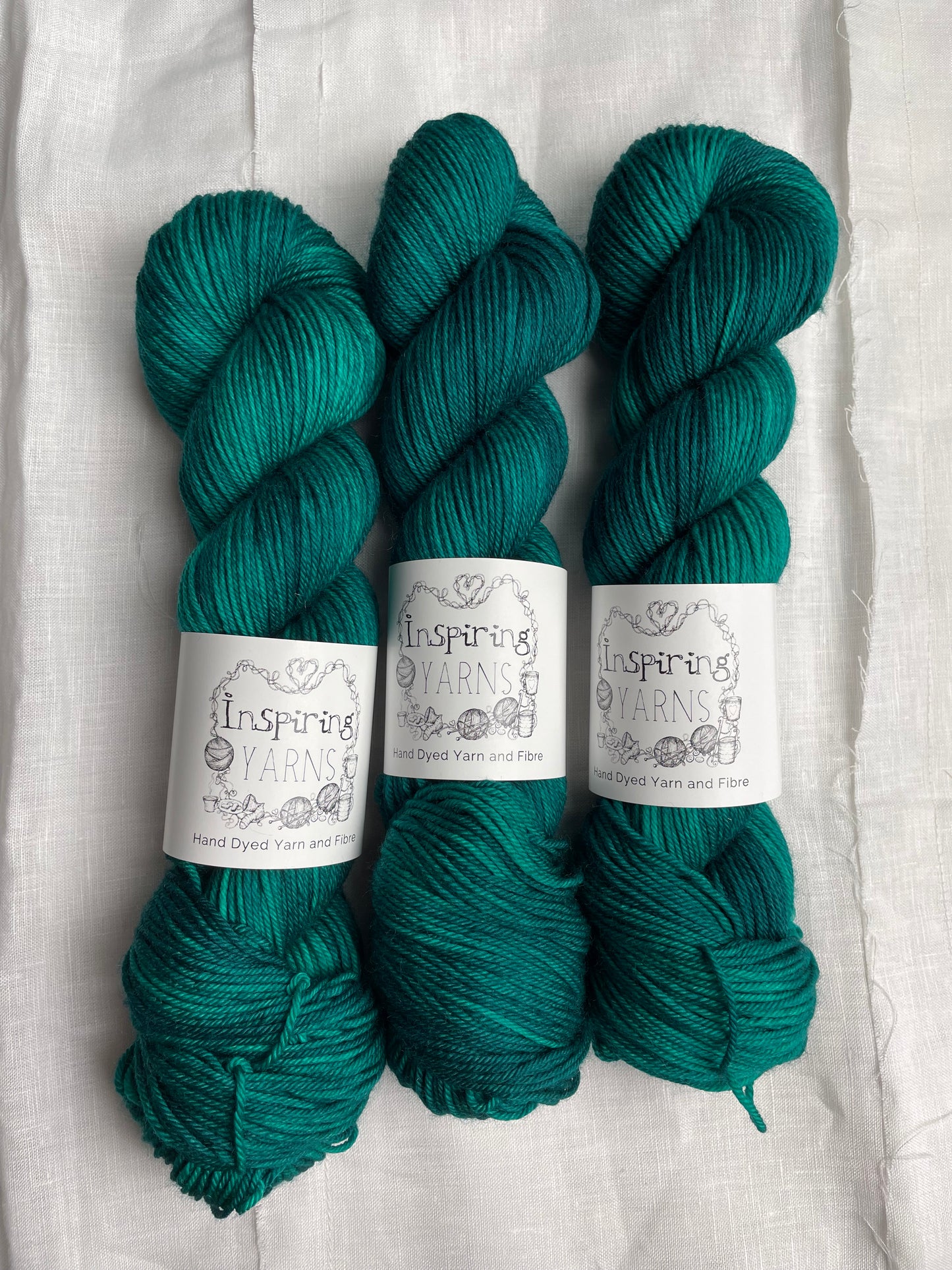 Three skeins of Merino Smooth DK yarn hand dyed in a tonal jade green colourway and labelled Inspiring Yarns on a white linen background.