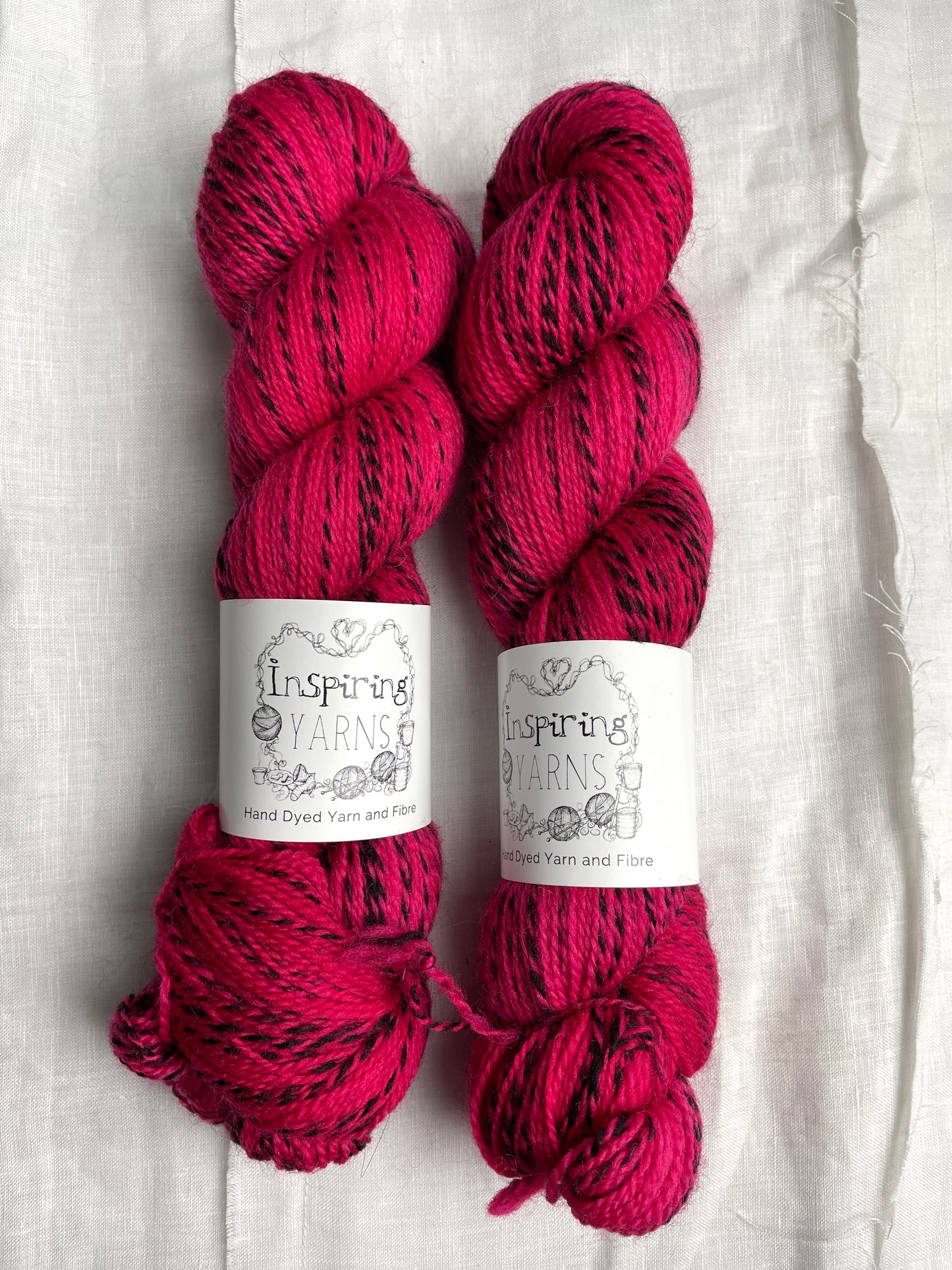 Two skeins of handyed Highland Zebra yarn in a bright cerise pink colourway. The yarn has black strands spun in to it. the skeins are labelled as Inspiring Yarns.