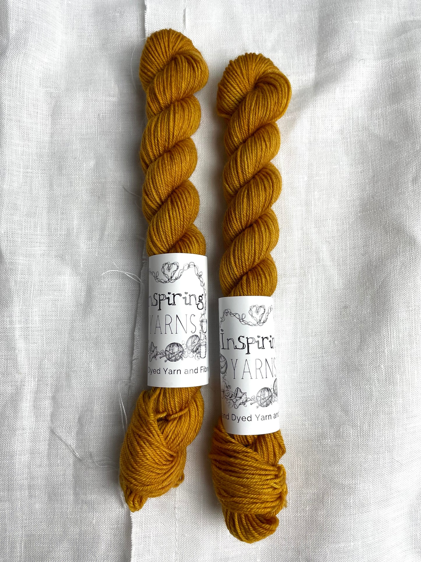 Two skeins of Keen As, a deep mustard tonal coloured Merino Smooth Sock, labelled Inspiring Yarns on a white linen background.