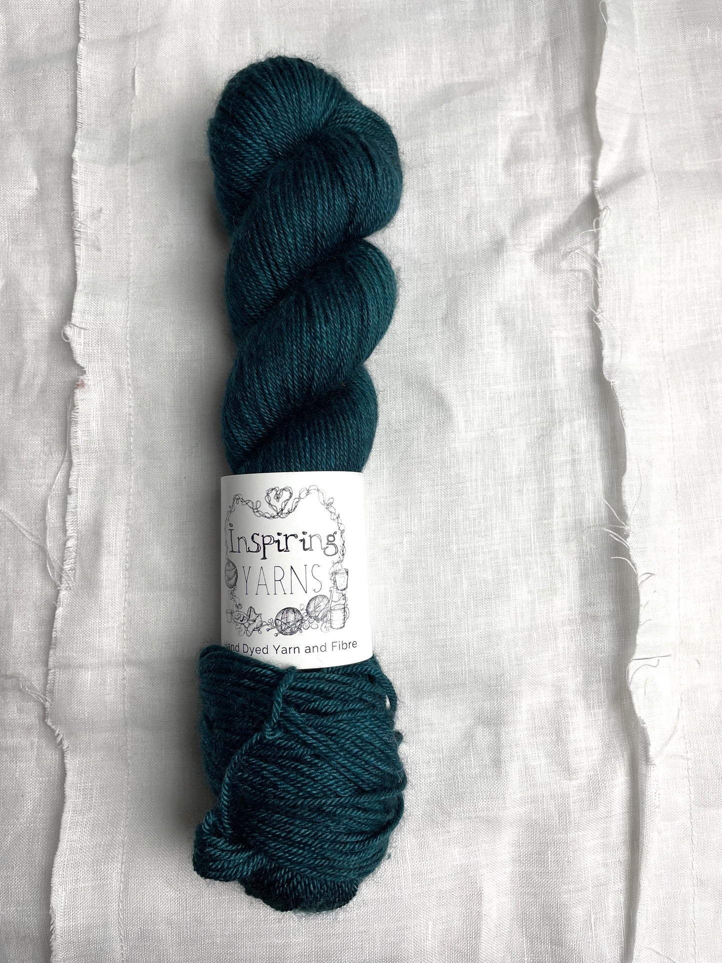 One skein of teal blue coloured yak silk dk yarn branded Inspiring Yarns on a white linen background.