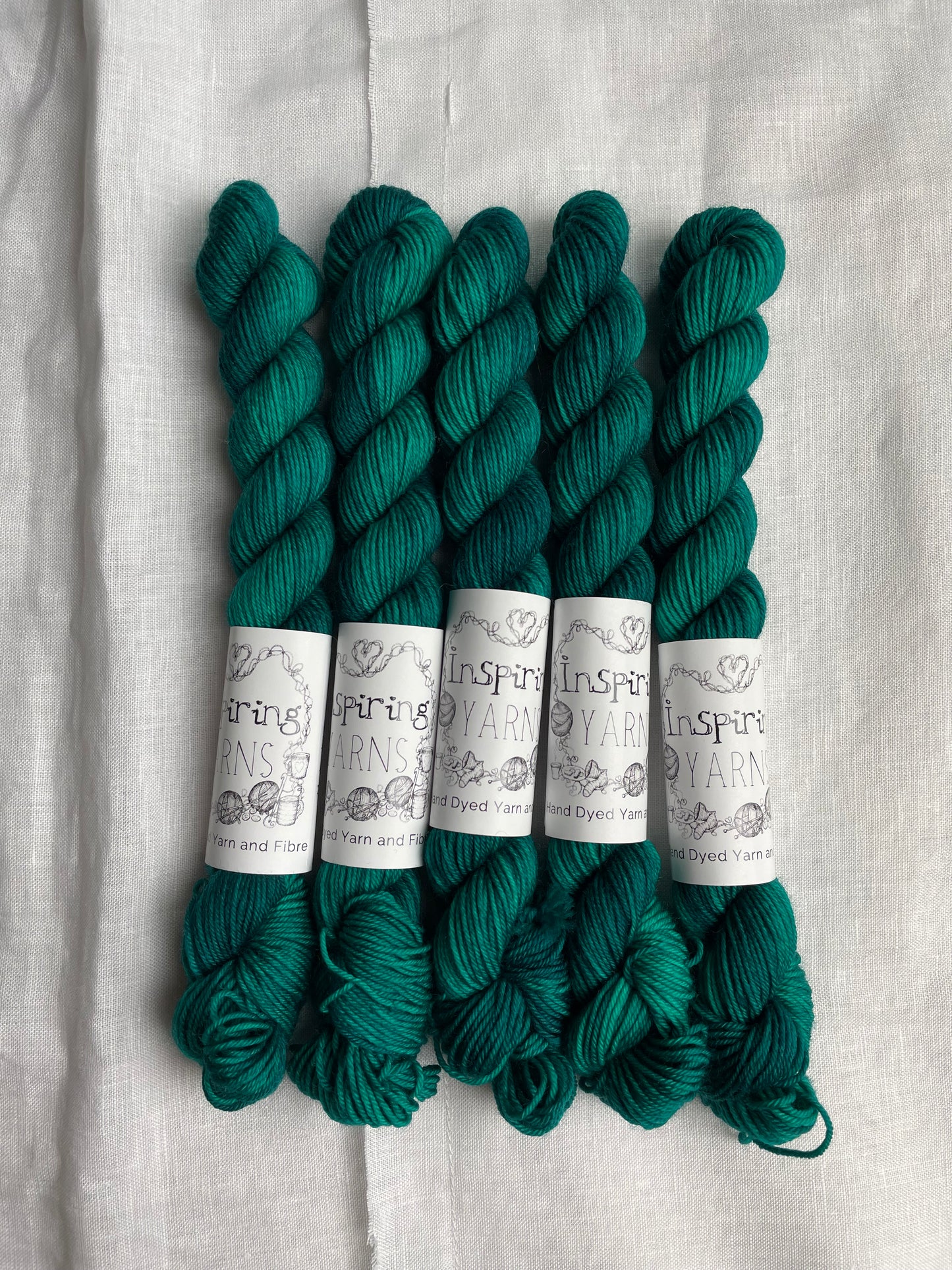 Five mini skeins of Merino Smooth Sock yarn hand dyed in a tonal jade green colourway and labelled Inspiring Yarns on a white linen background.