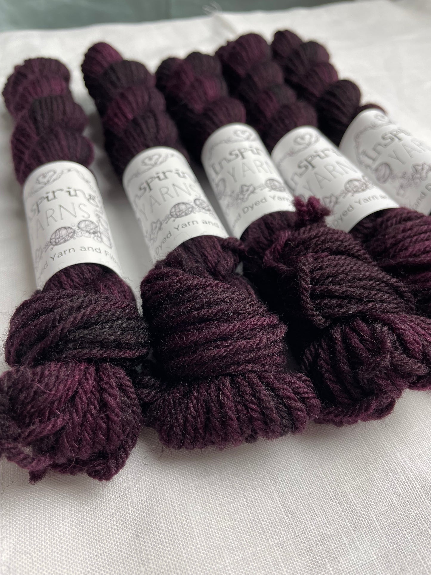 Five skeins of 20 gram dark burgundy coloured Highland 4Ply labelled Inspiring Yarns on a white linen background.