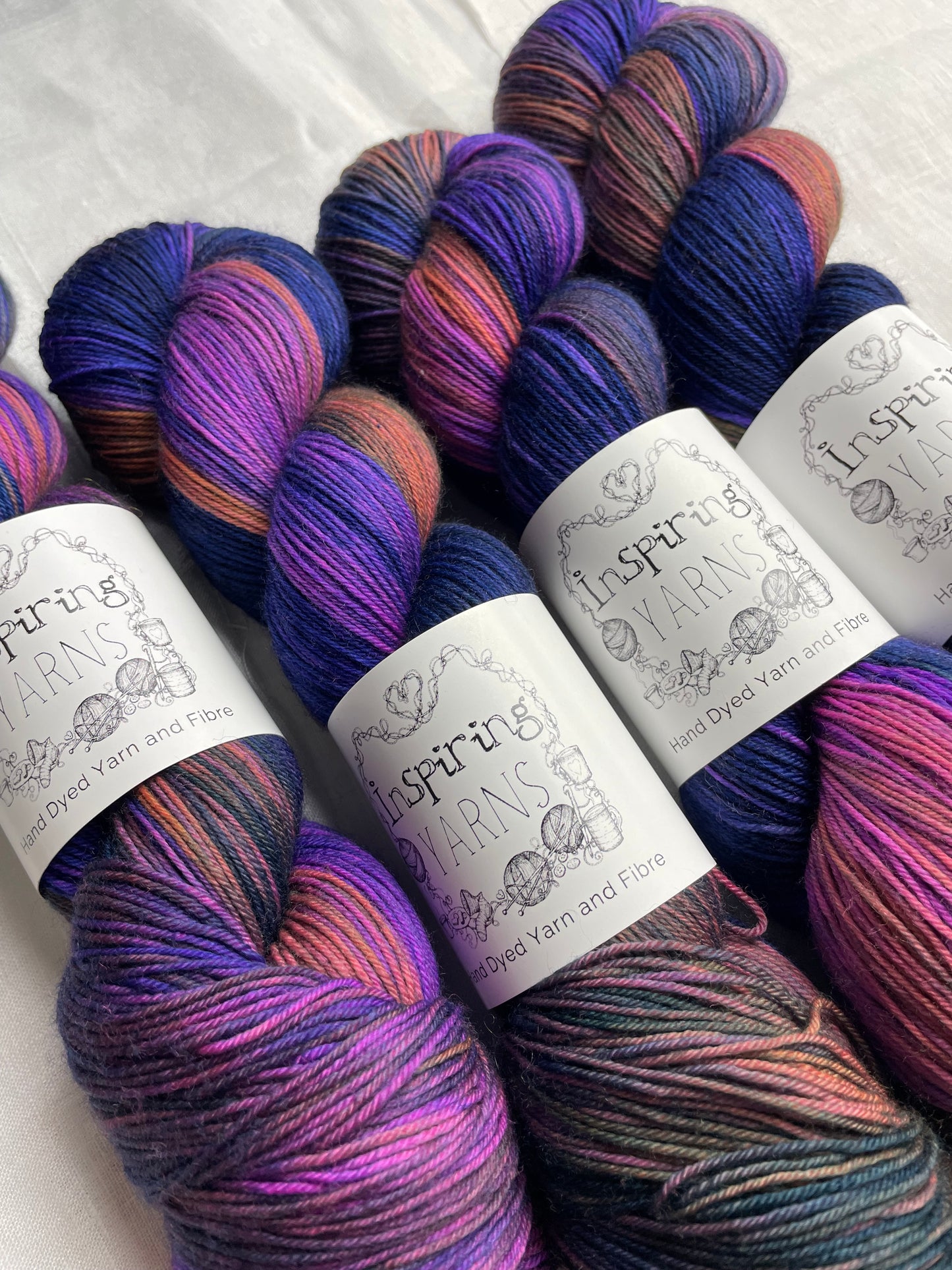 Close up of four variegated blue and multi coloured skeins of Merino Smooth Sock yarn branded Inspiring Yarns on a white linen fabric background.