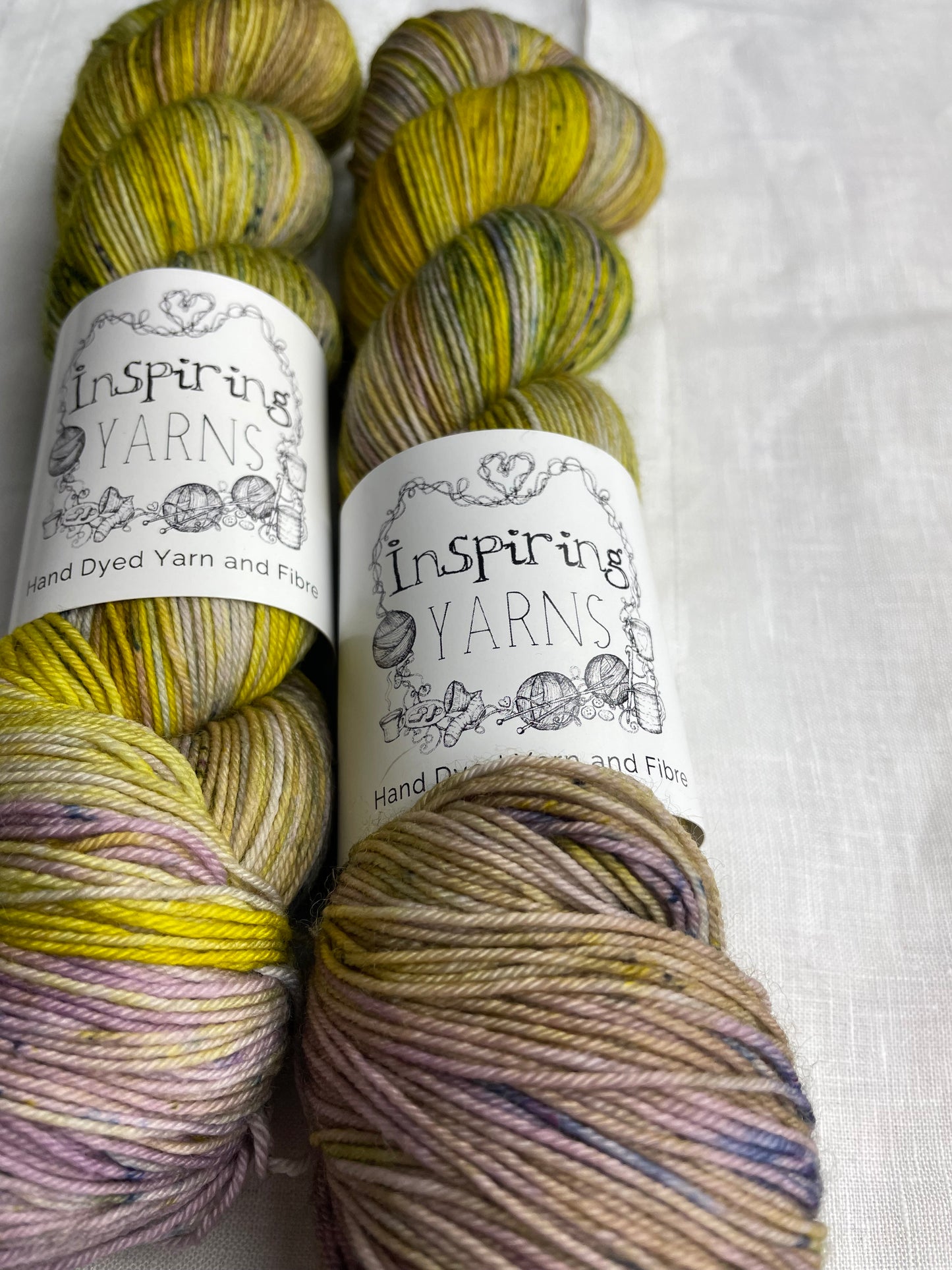 Close up of two acid yellow and lilac coloured skeins of Merino Smooth Sock yarn branded Inspiring Yarns on a white linen fabric background.