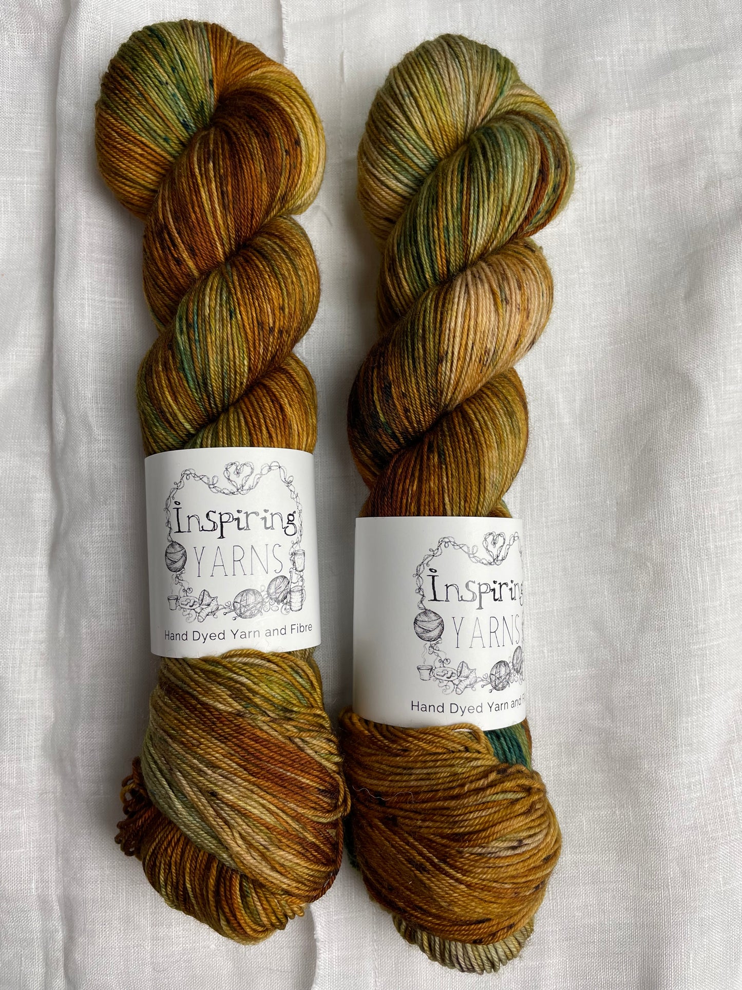 Two skeins of Get Cosy, variegated shades of tan with teal speckles coloured Merino Smooth Sock, labelled Inspiring Yarns on a white linen background.