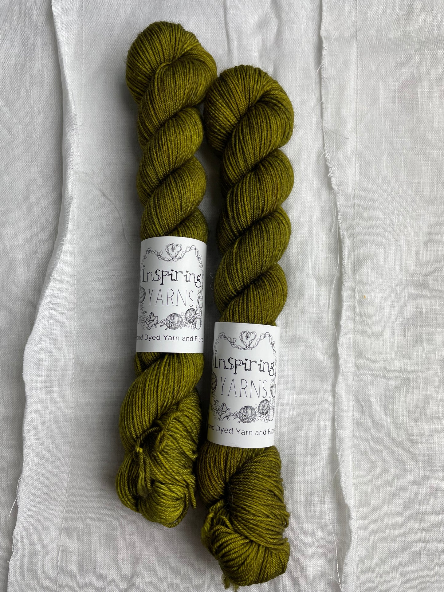 Two dark olive green coloured skeins of hand dyed Merino Smooth Sock yarn branded Inspiring Yarns on white linen fabric.