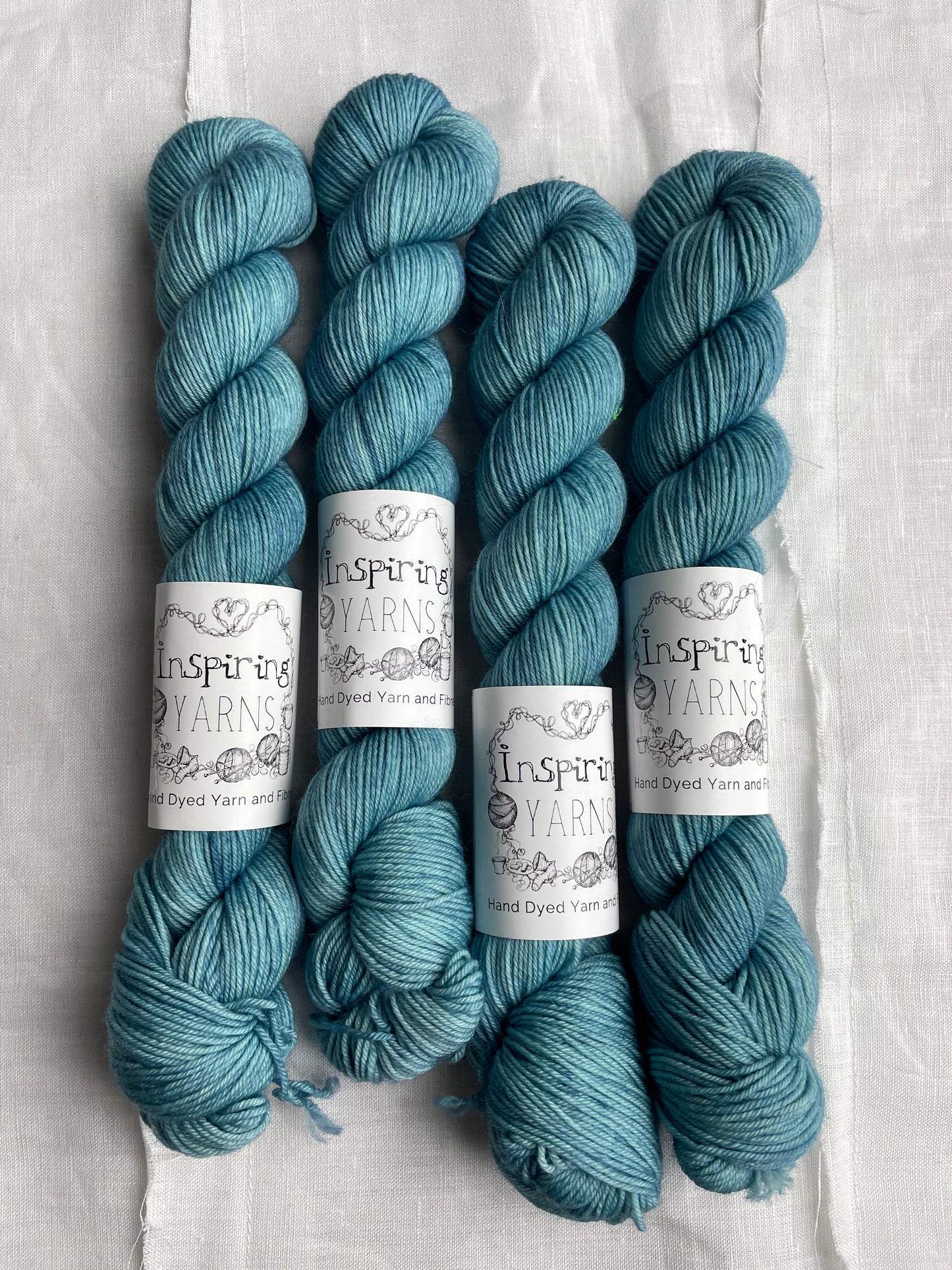 Four tonal soft teal coloured skeins of hand dyed Merino Smooth Sock yarn branded Inspiring Yarns on white linen fabric.