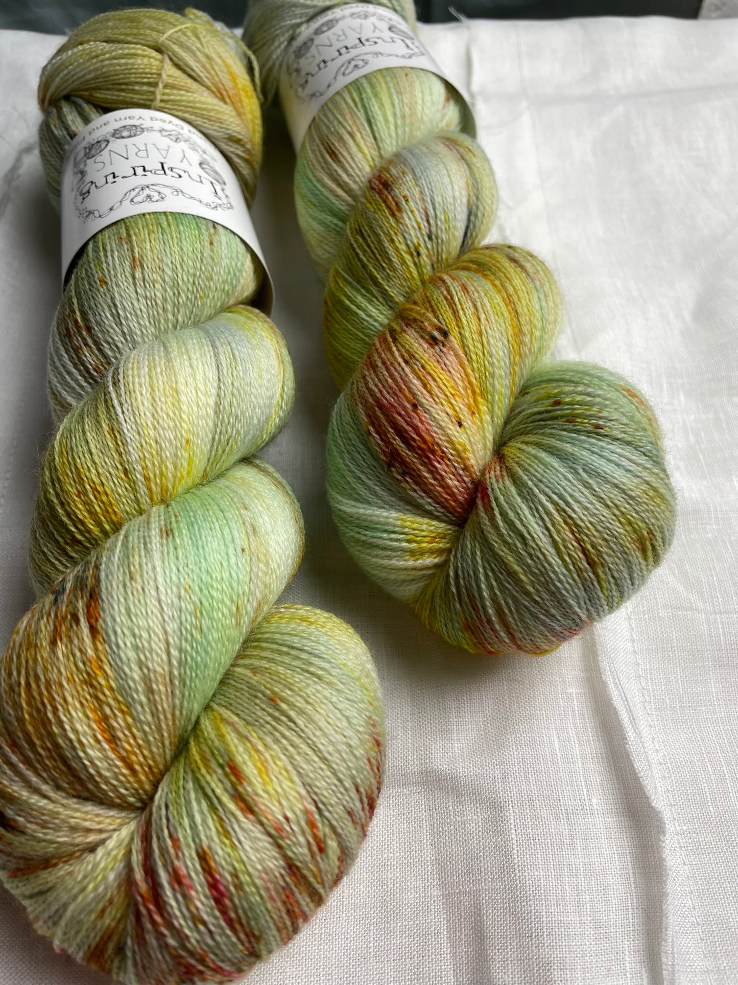Close up of two skeins of speckled silver grey and mint coloured hand dyed Merino Lace yarn branded Inspiring Yarns on a white linen cloth.
