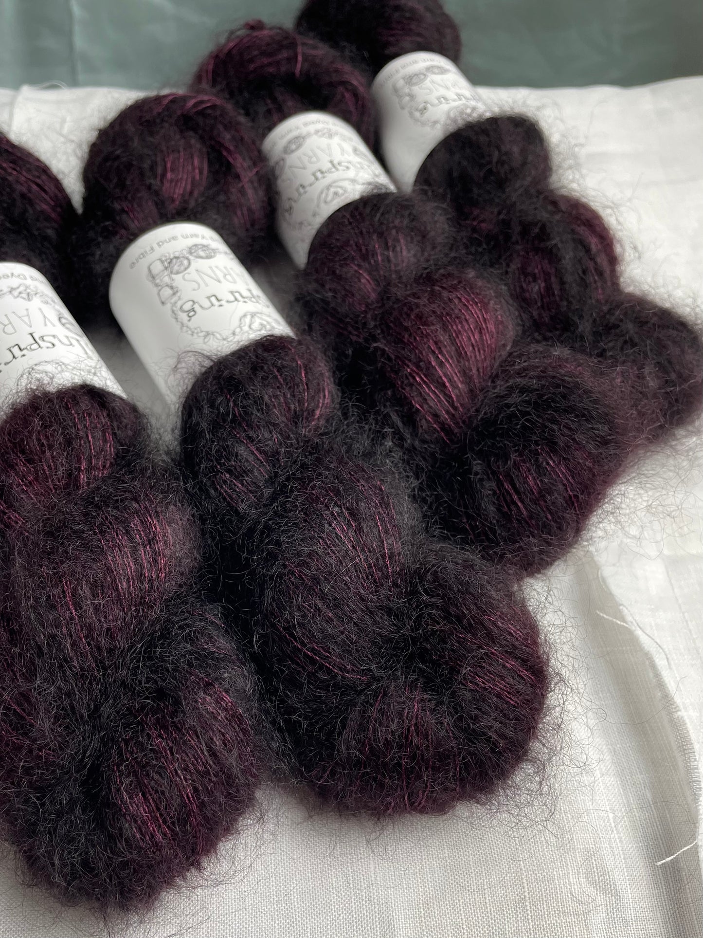 Four skeins of dark burgundy coloured Kid Mohair Silk Lace labelled Inspiring Yarns on a white linen background.