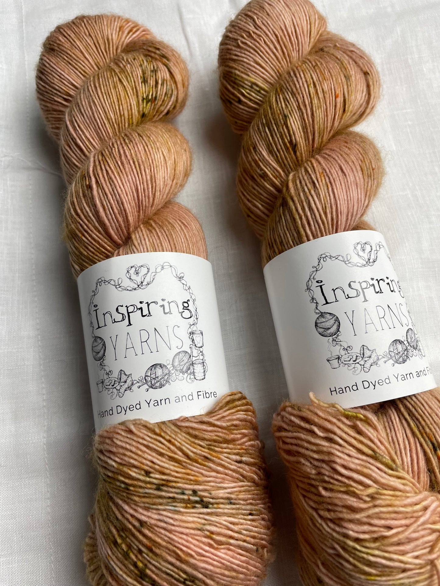 Close up of two skeins of speckled soft terracotta coloured hand dyed Merino Singles yarn labelled Inspiring Yarns on a white linen background.