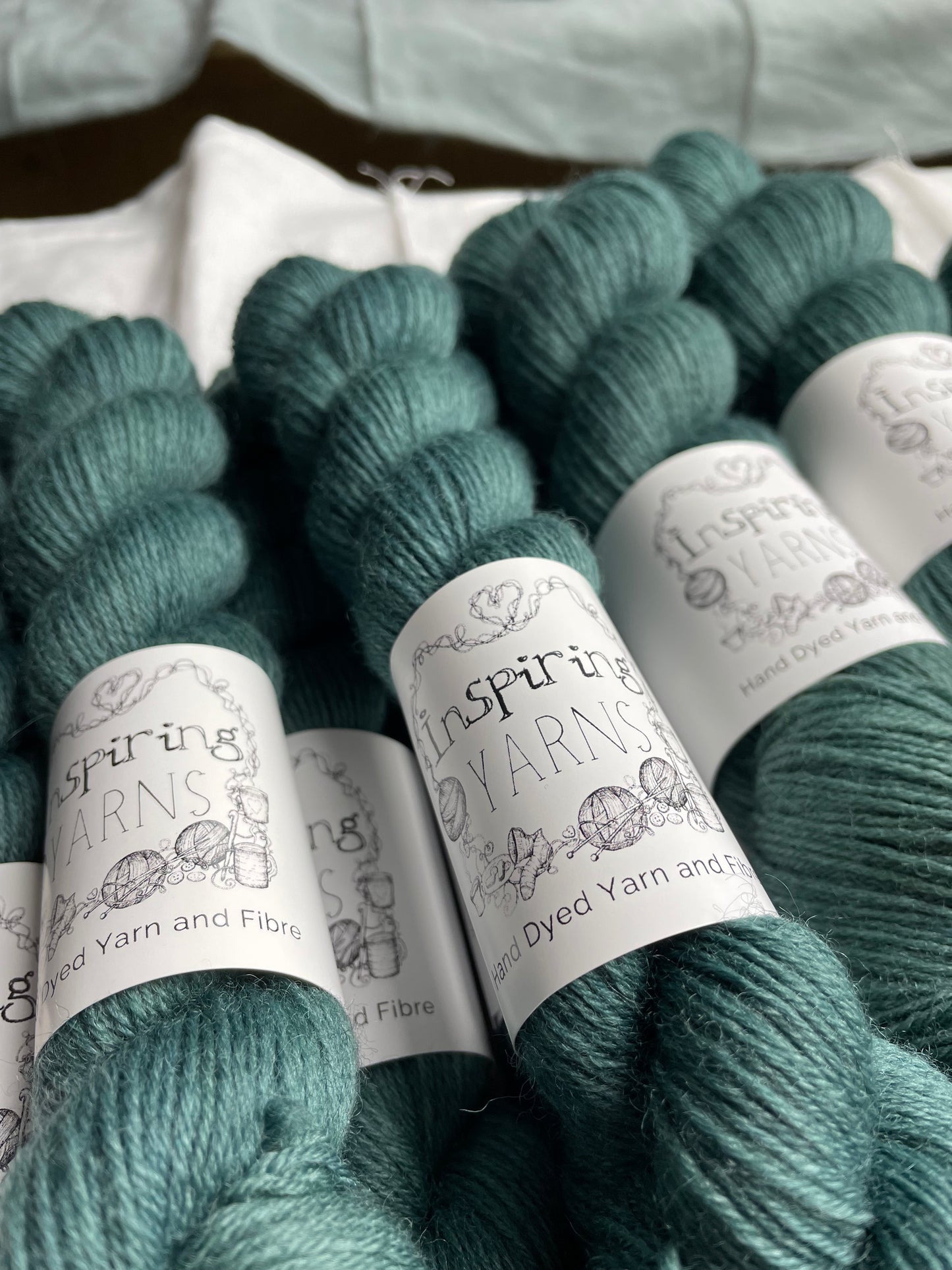 Multiple skeins of a tonal dark teal Highland 4Ply yarn branded Inspiring Yarns on a white linen background.