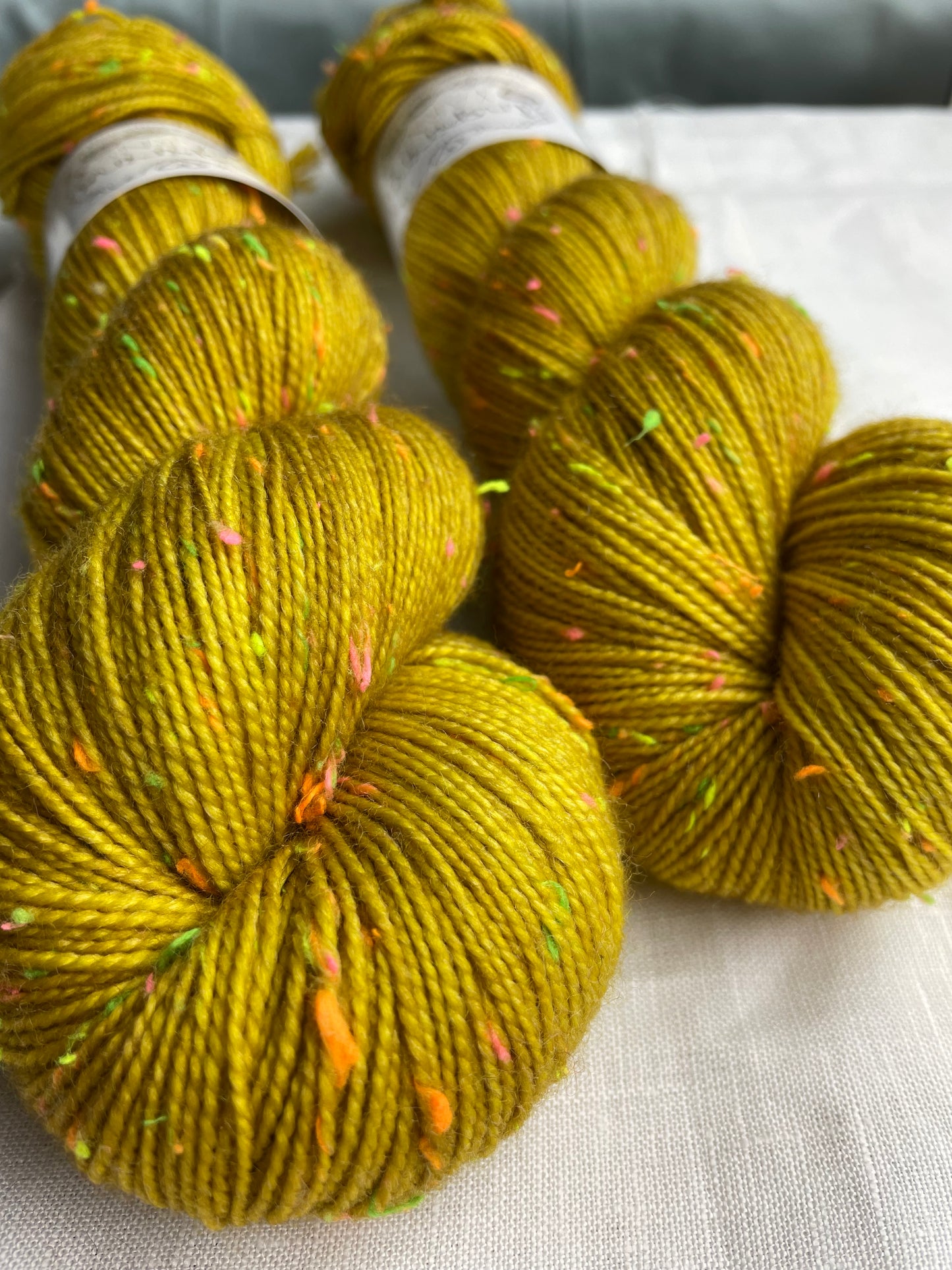 Close up of two skeins of tonal golden ochre coloured hand dyed Disco Donegal yarn branded Inspiring Yarns on a white linen background. The yarn has multi coloured neon neps.