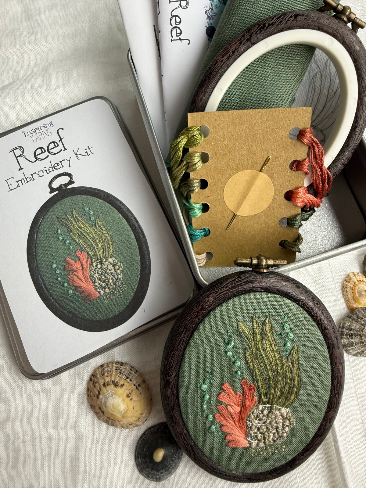 A coral reef-themed embroidery kit shown in a styled flat-lay. The photograph shows the completed embroidery in a dark oval frame, a metal tin filled with thread, needle, frame, fabric and instructions, and the tin lid with a picture of the design, photographed with sea shells on a white background.