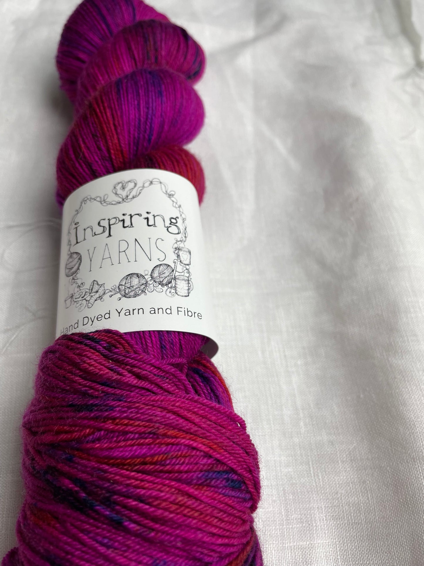 A skein of fuchsia pink speckled Merino Smooth Sock labelled InspiringYarns lies on a white linen background.