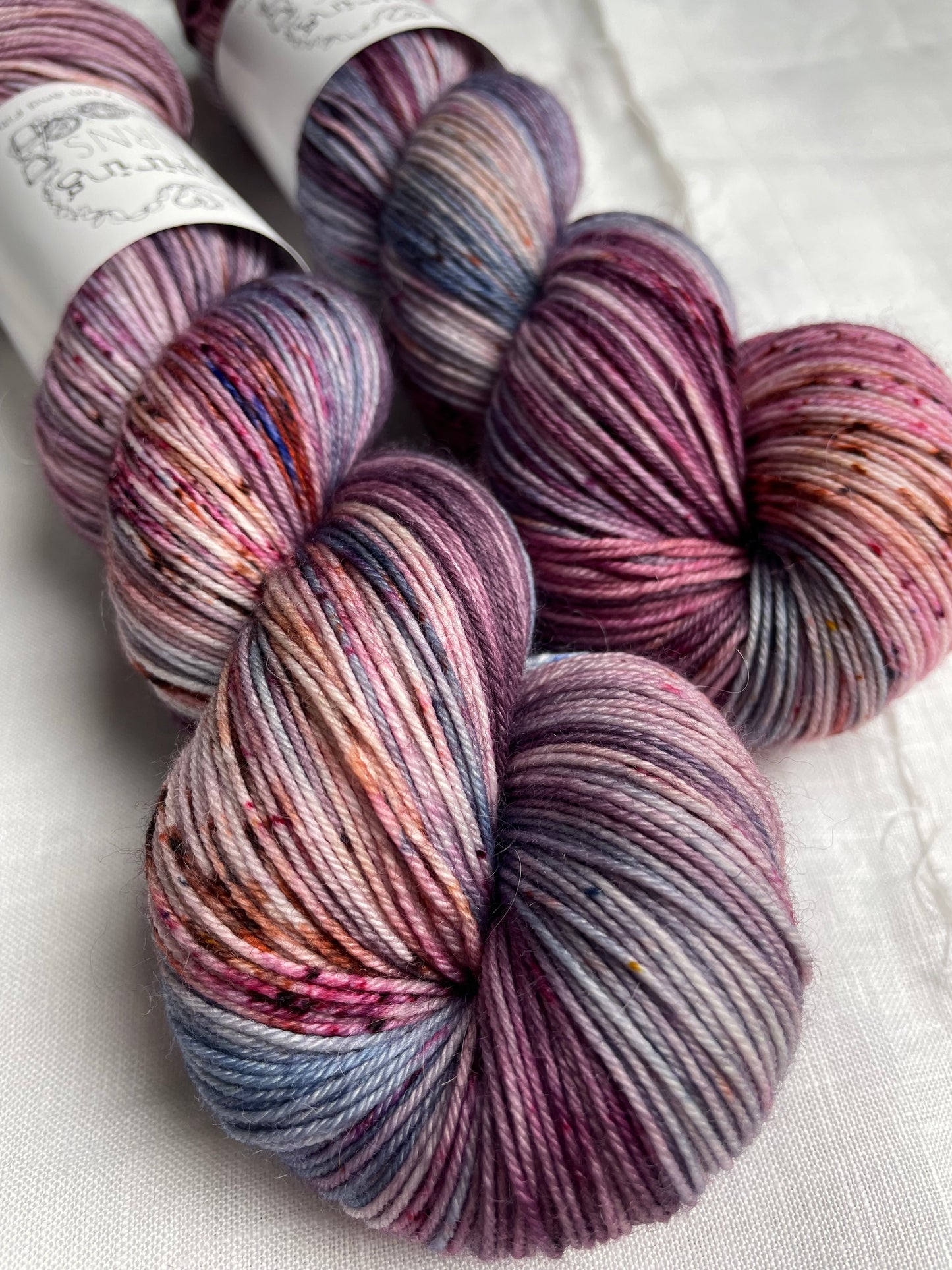 Two skeins of merino smooth sock yarn in shades of pink and blue labelled Inspiring Yarns on a white linen background.