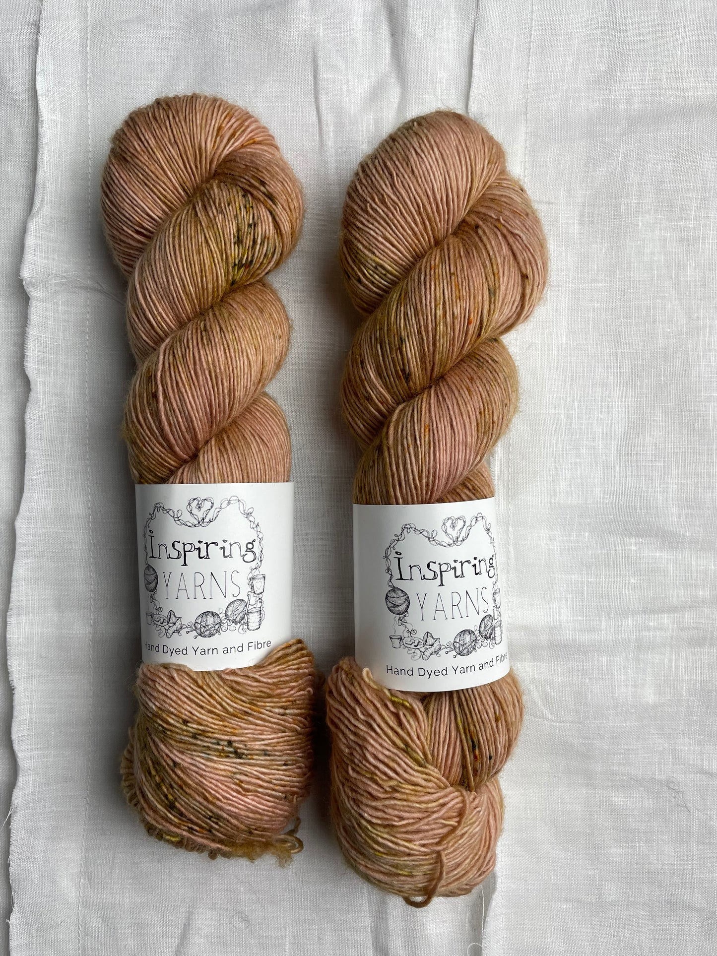 Two skeins of speckled soft terracotta coloured hand dyed Merino Singles yarn labelled Inspiring Yarns on a white linen background.