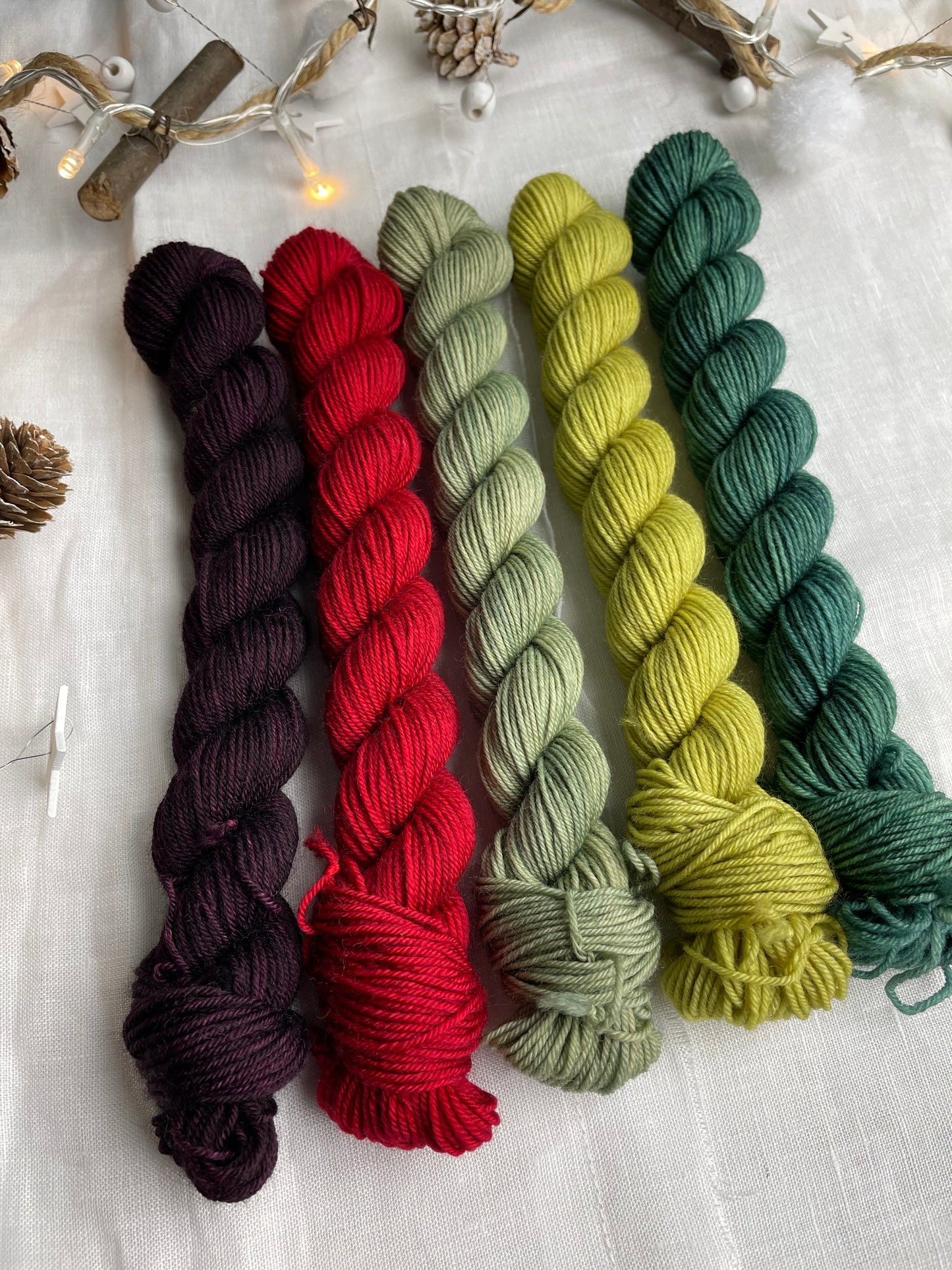 A set of five hand dyed yarn mini skeins on Merino Smooth Sock Yarn in red and green tones labelled Inspiring Yarns. The background is white linen and some Christmas lights with pinecones, twigs and stars.