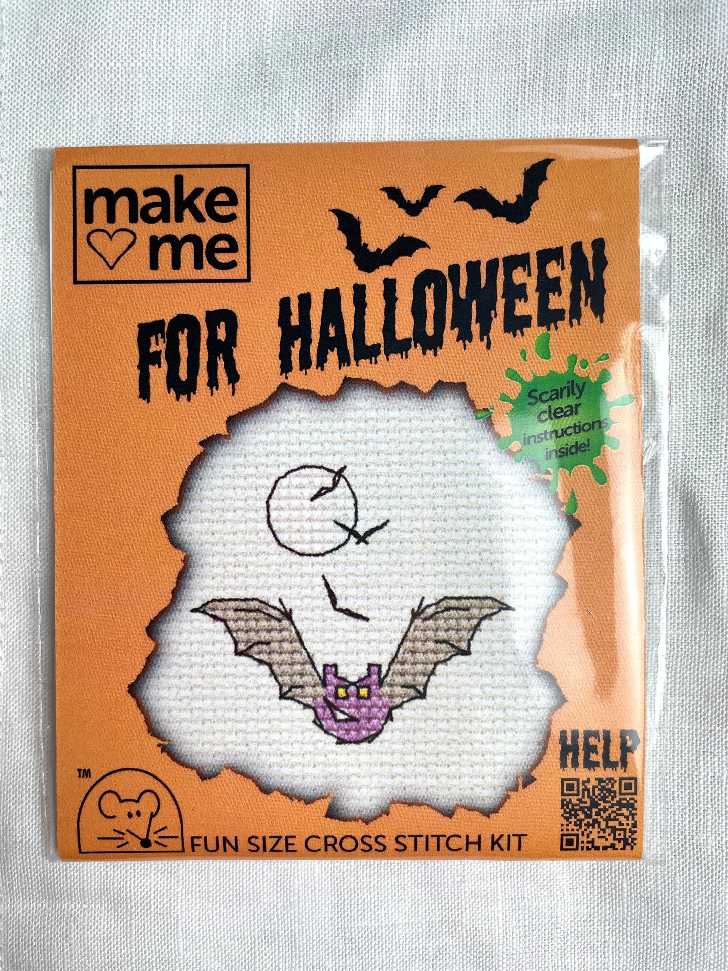 A mini crosstitch kit for a purple bat and smaller bats flying in front of the moon photographed on a white linen background. It is branded Mouseloft Make Me For Halloween in orange packaging.