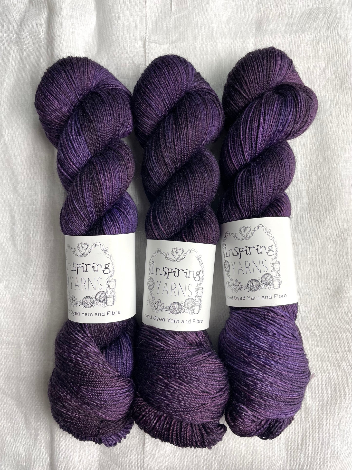 Three skeins of merino smooth sock in a tonal dark purple colour labelled Inspiring Yarns on a white linen background.