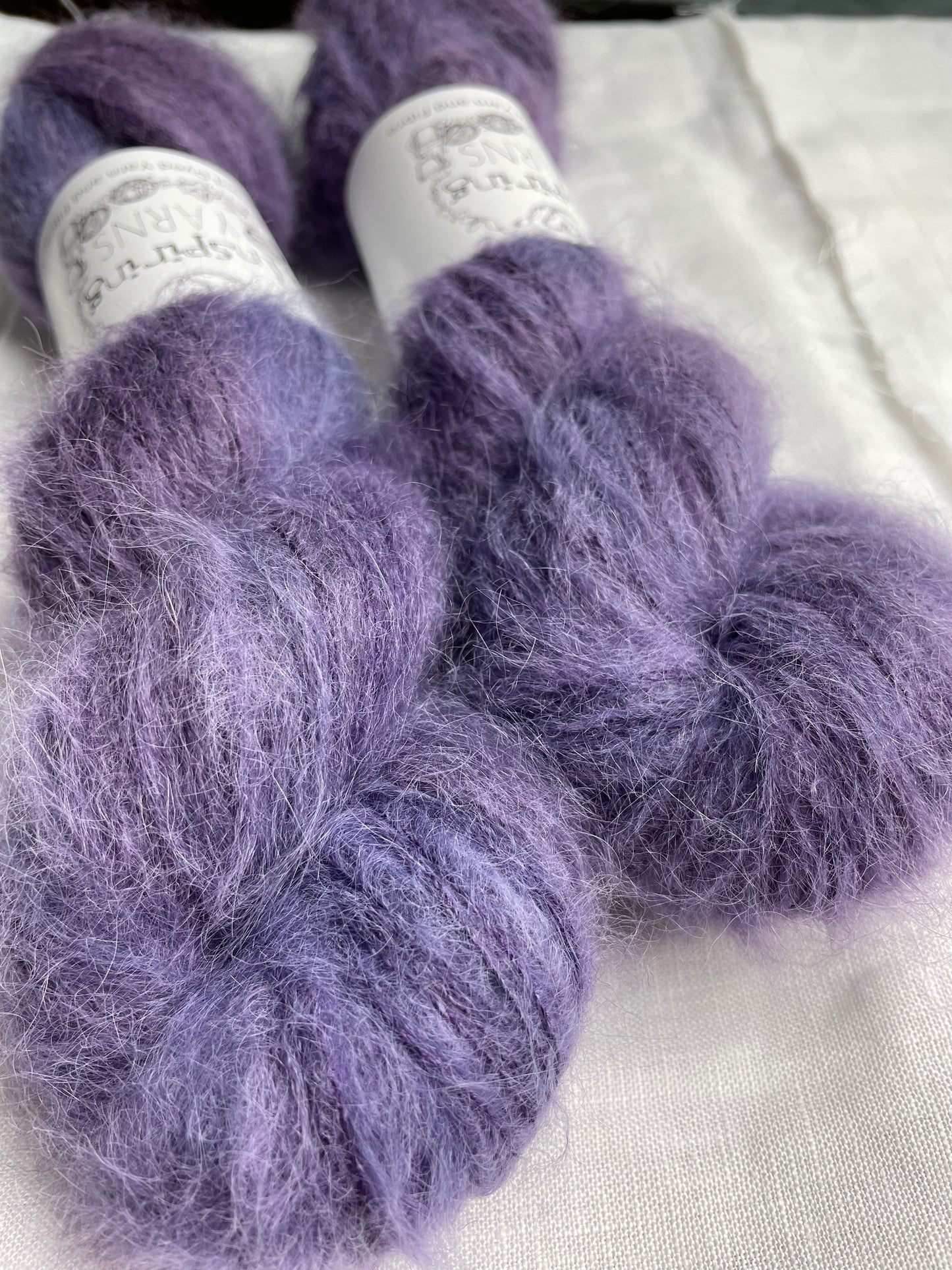 Two skeins of brushed suri silk yarn in a tonal dark purple colour labelled Inspiring Yarns on a white linen background.