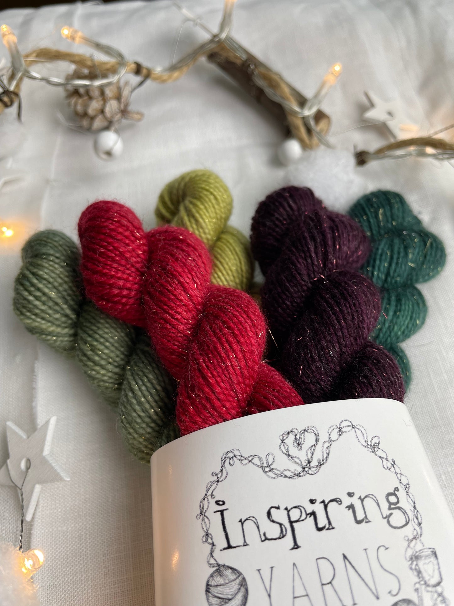 A close up of a set of five hand dyed yarn mini skeins on Sparkle Sock Yarn in red and green tones labelled Inspiring Yarns. The background is white linen and some Christmas lights with pinecones, twigs and stars.