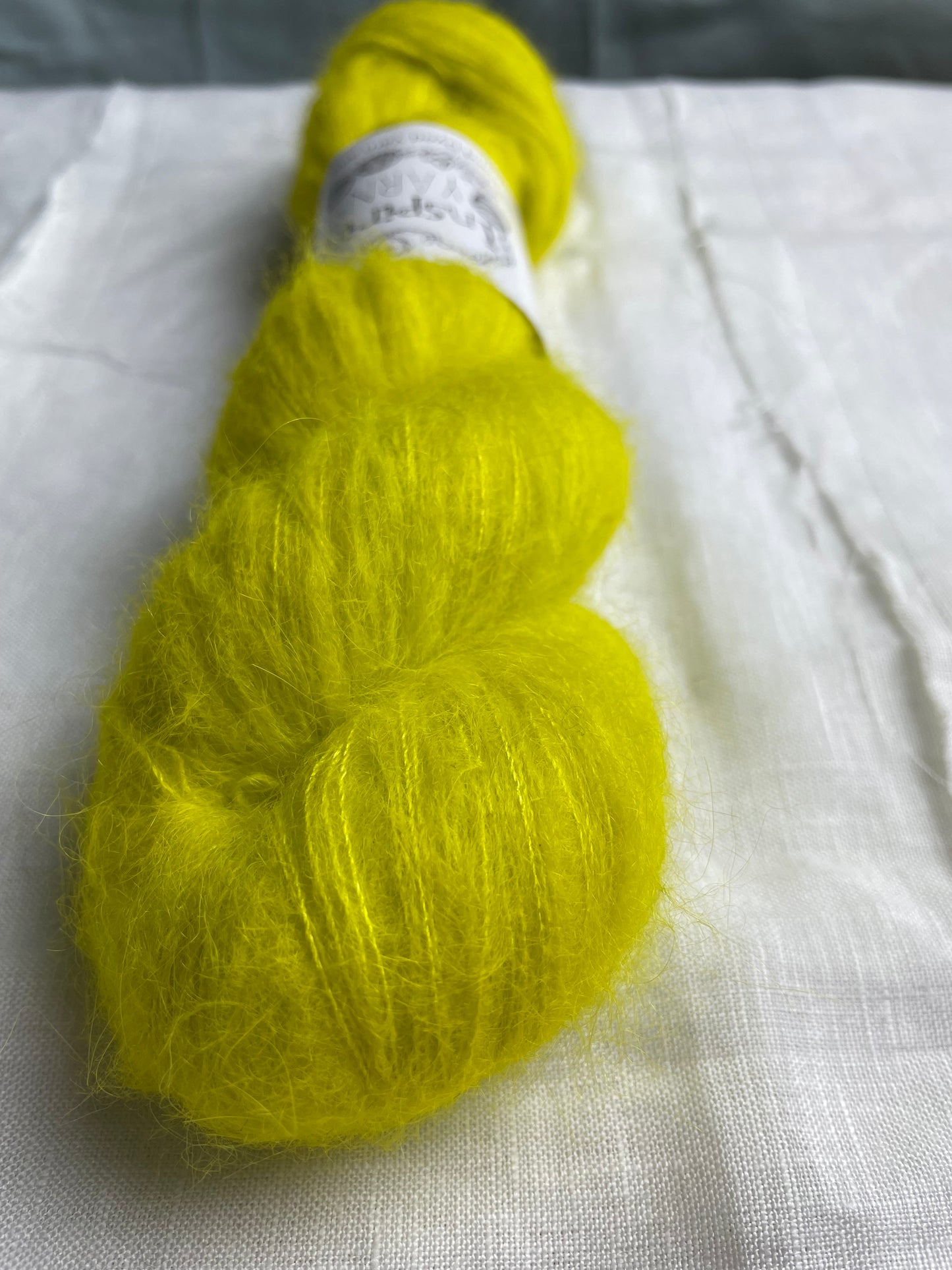 Close up of a skein of tonal acid yellow coloured hand dyed fluffy Alpaca Silk Lace yarn labelled Inspiring Yarns on a white linen background.