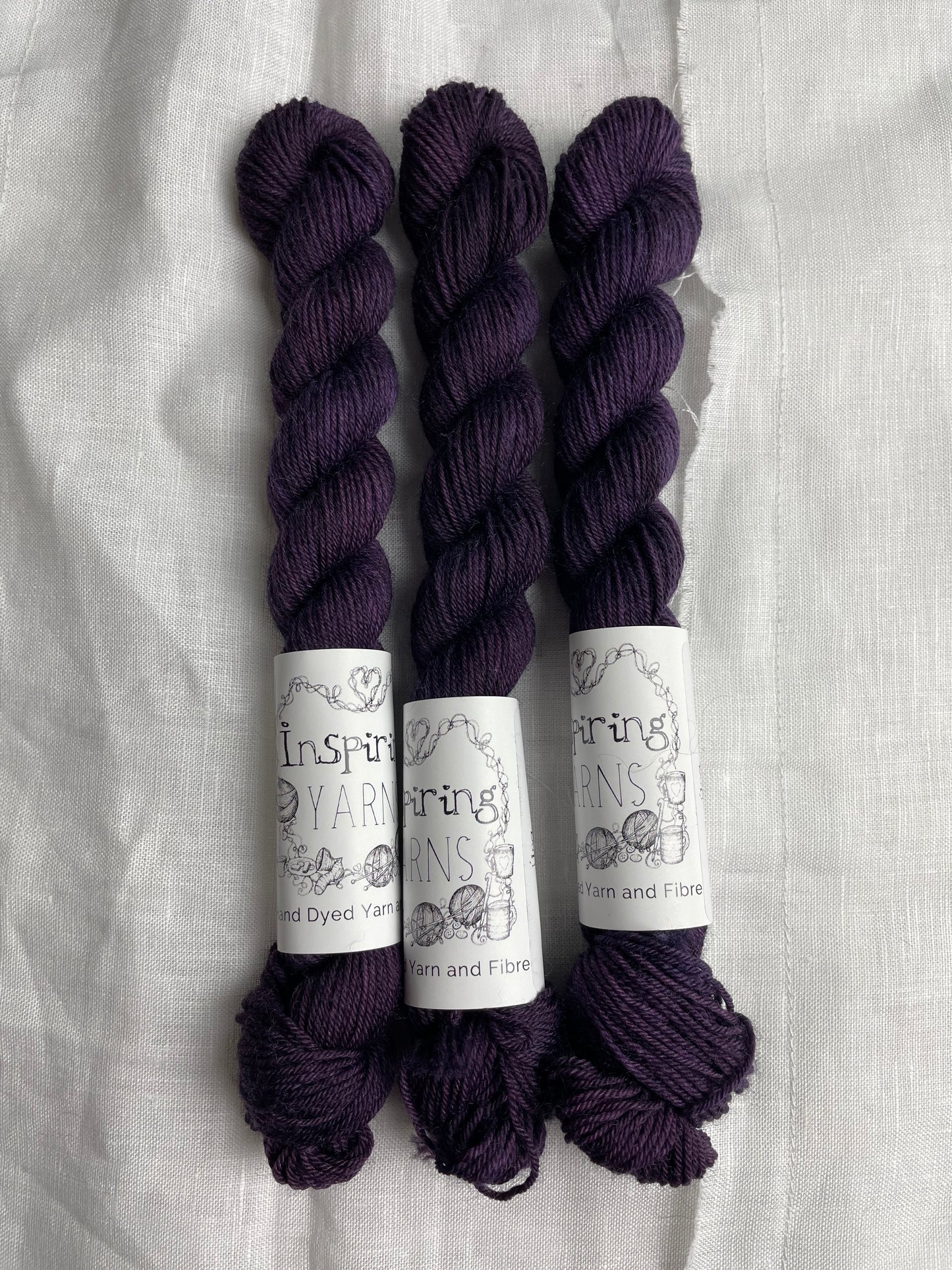 Three mini skeins of dark purple Merino Smooth Sock yarn labelled Inspiring Yarns on a white linen background.