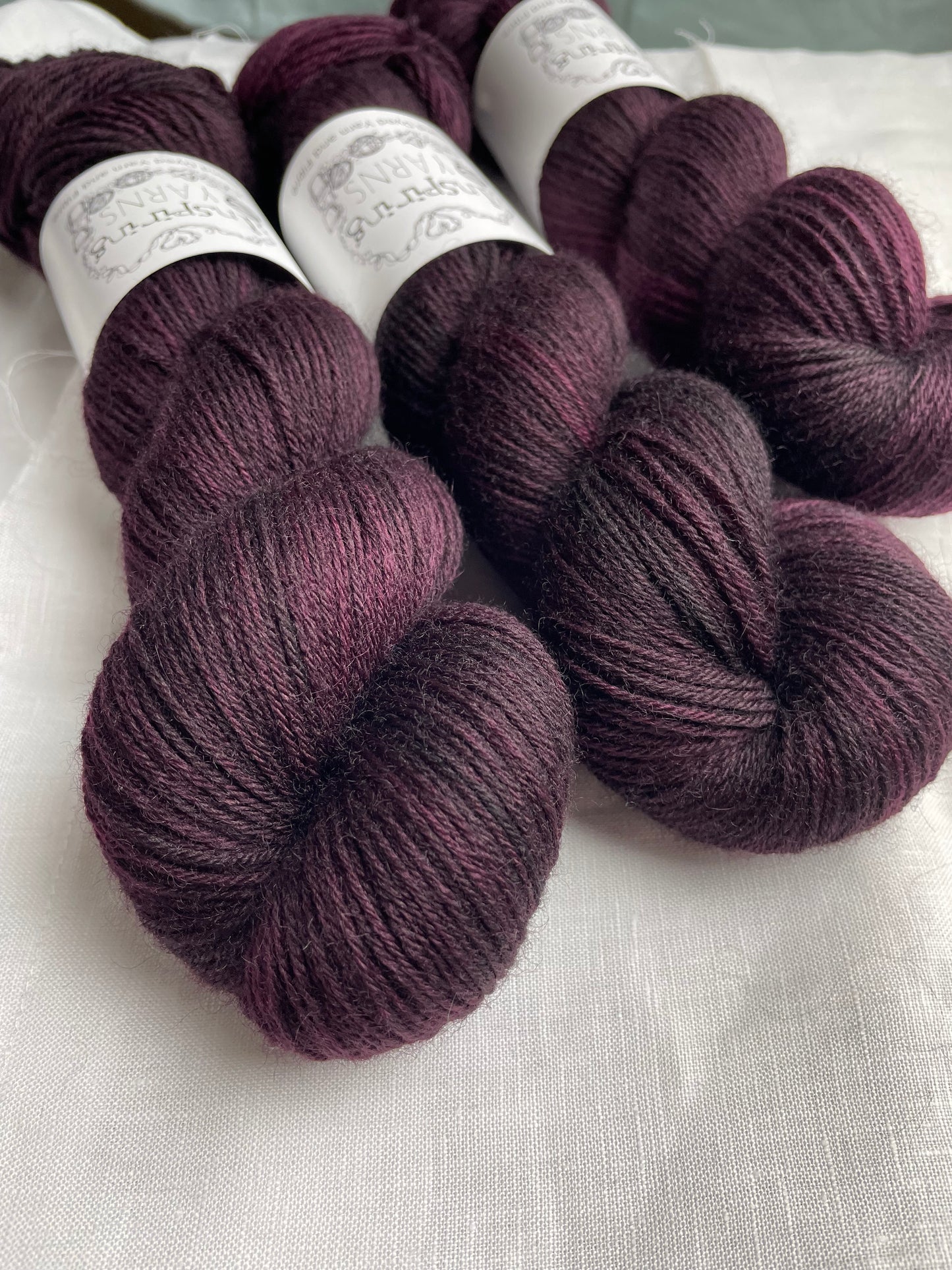 Three skeins of dark burgundy coloured Highland 4Ply labelled Inspiring Yarns on a white linen background.