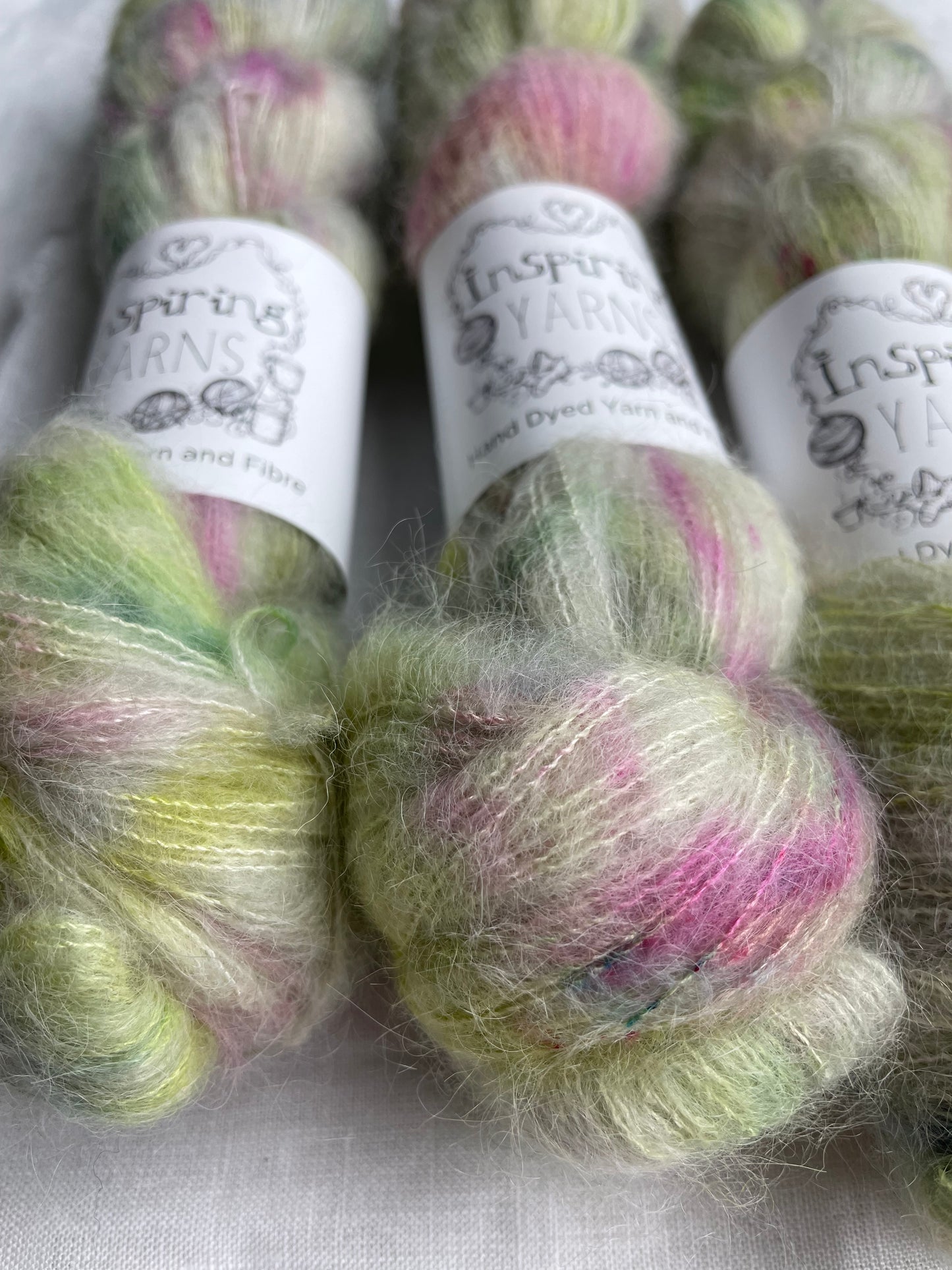 Close up of three skeins of green and pink speckled silver grey coloured hand dyed fluffy Alpaca Silk Lace yarn labelled Inspiring Yarns on a white linen background.