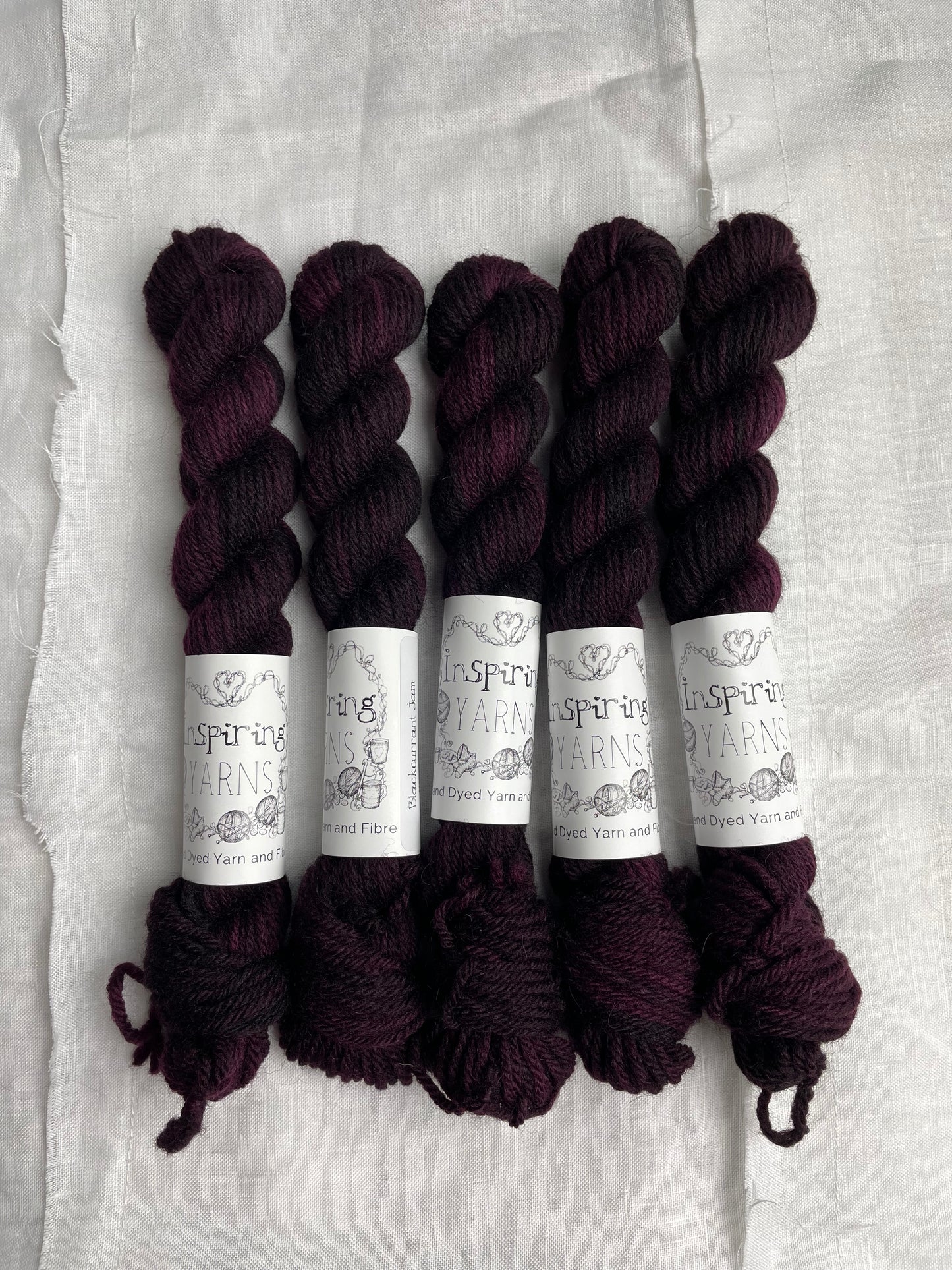 Five skeins of 20 gram dark burgundy coloured Highland 4Ply labelled Inspiring Yarns on a white linen background.
