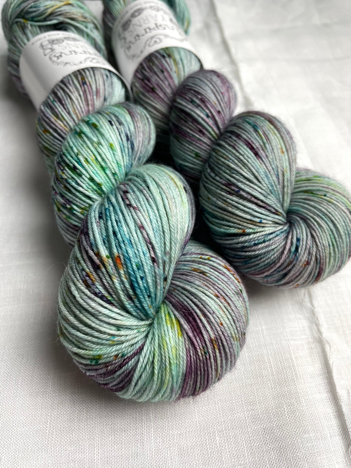 Two skeins of merino smooth sock yarn in a light teal colour with coloured speckles labelled Inspiring Yarns on a white linen background.