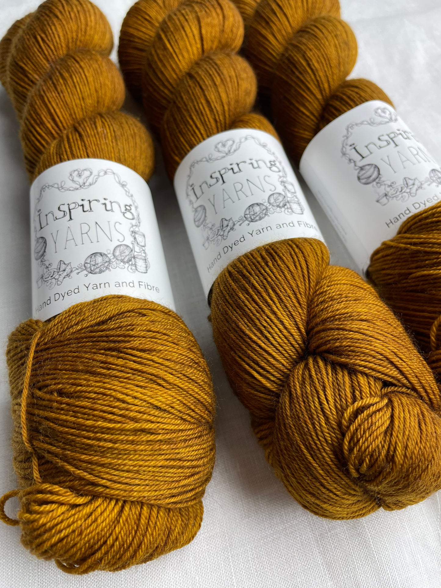 Close up of three tonal toffee brown coloured skeins of Merino Smooth Sock yarn branded Inspiring Yarns on a white linen fabric background.