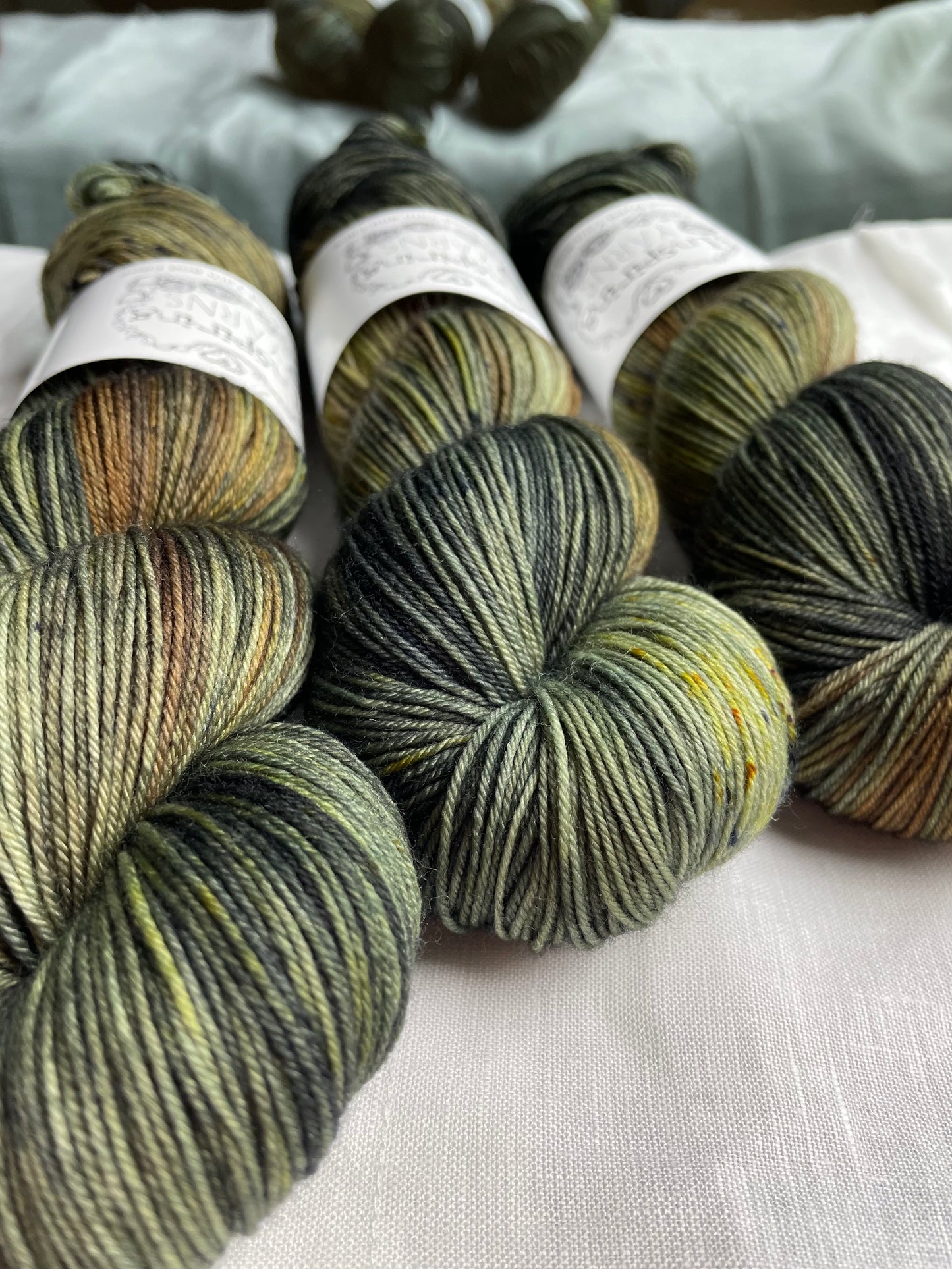 up close image of three skeins of green yarn with inspiring yans labels and a reflective back ground and a fabric covered suface.