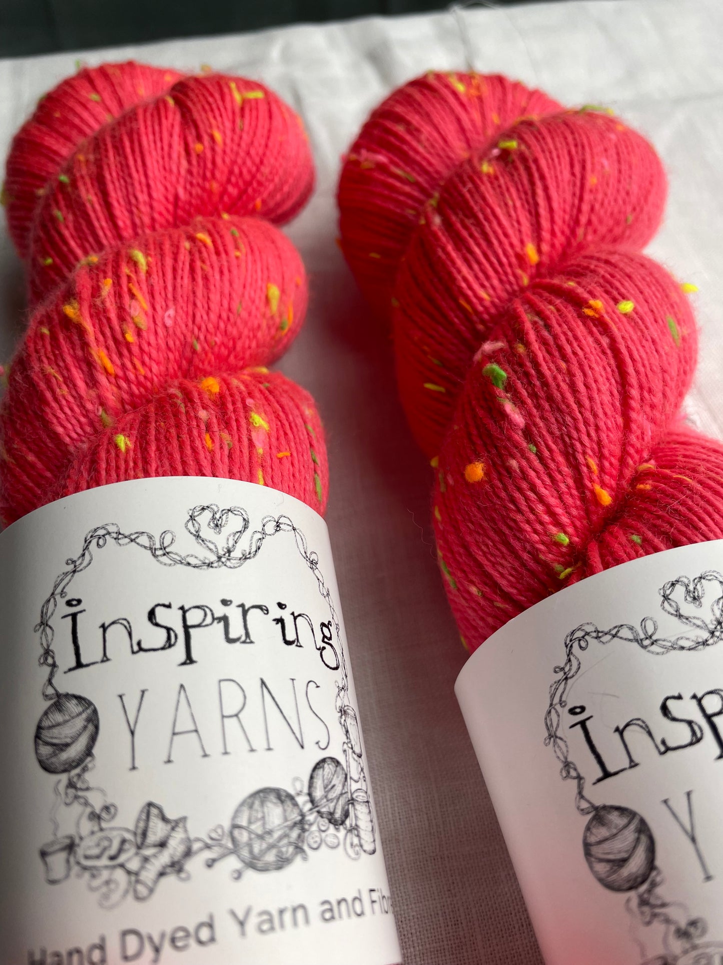 Close up of two skeins of tonal dark neon coral coloured hand dyed Disco Donegal yarn branded Inspiring Yarns on a white linen background. The yarn has multi coloured neon neps.