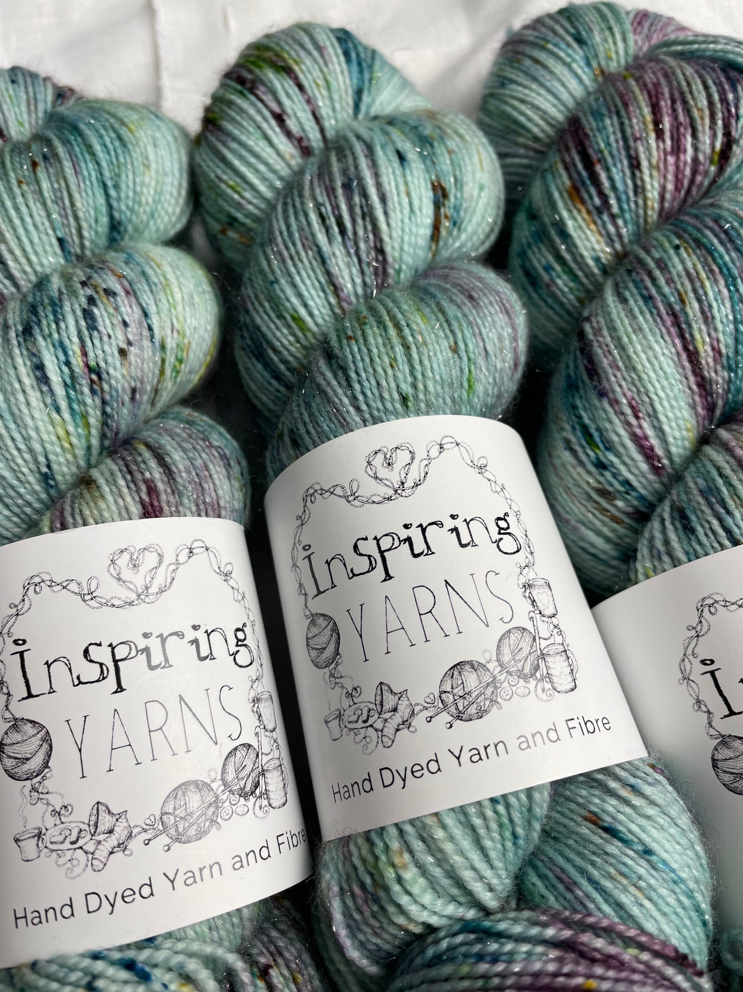 Three skeins of teal blue speckled Silver Sparkle Sock yarn labelled Inspiring Yarns on a white linen background.