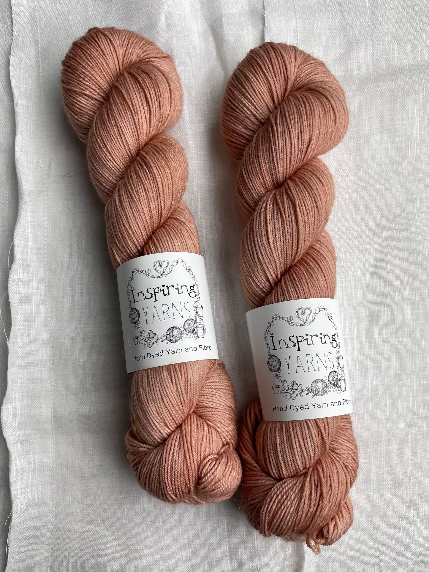Two dusky peach coloured skeins of Merino Smooth Sock yarn branded Inspiring Yarns on a white linen fabric background.